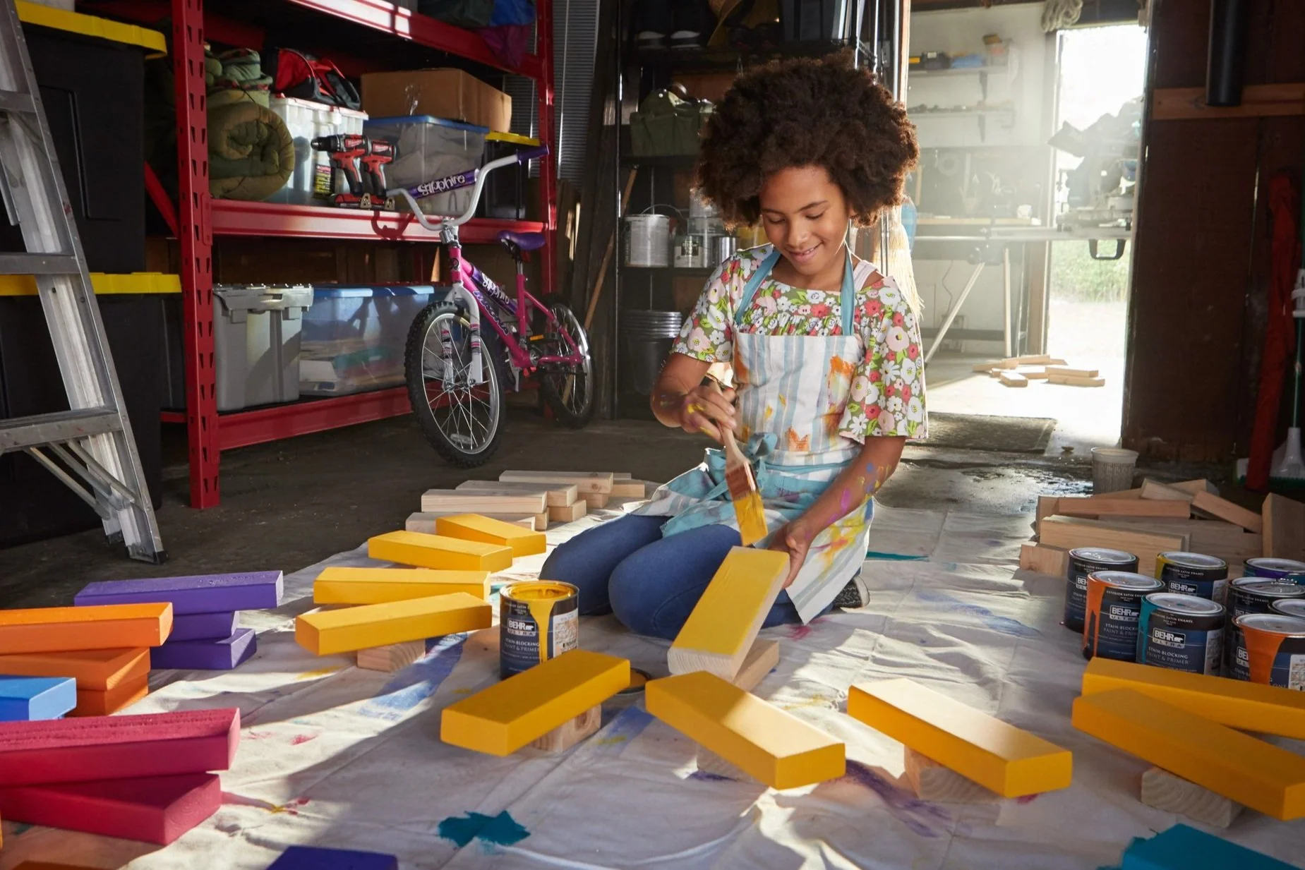 A young girl painting wooden blocks yellow in a garage, surrounded by paint cans and colorful wooden pieces, with a bicycle and storage shelves in the background.