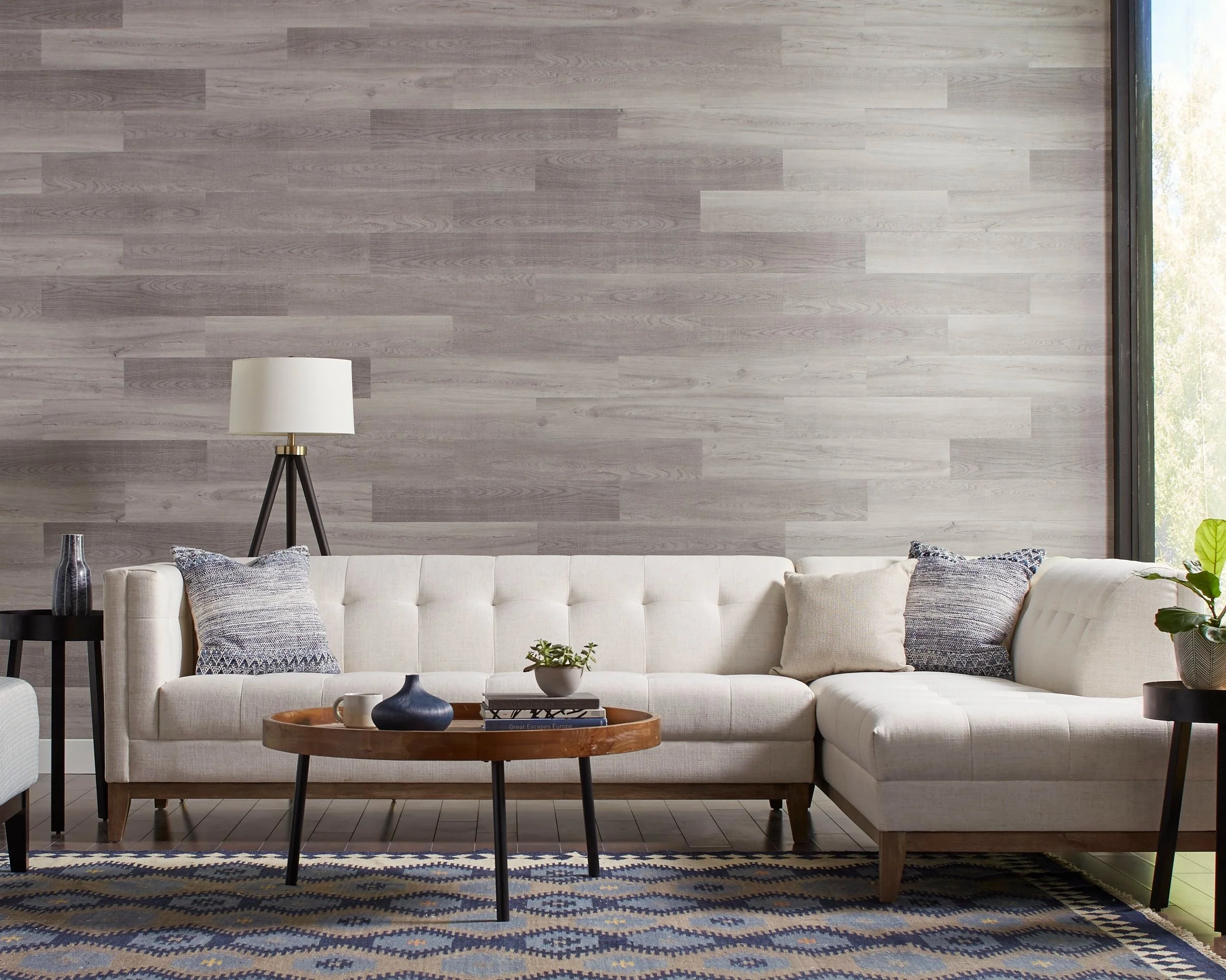 Living room with a white sectional sofa, blue and beige pillows, a wooden coffee table with decorative vases, a floor lamp, side tables with plants, a window with natural light, and a blue patterned rug.