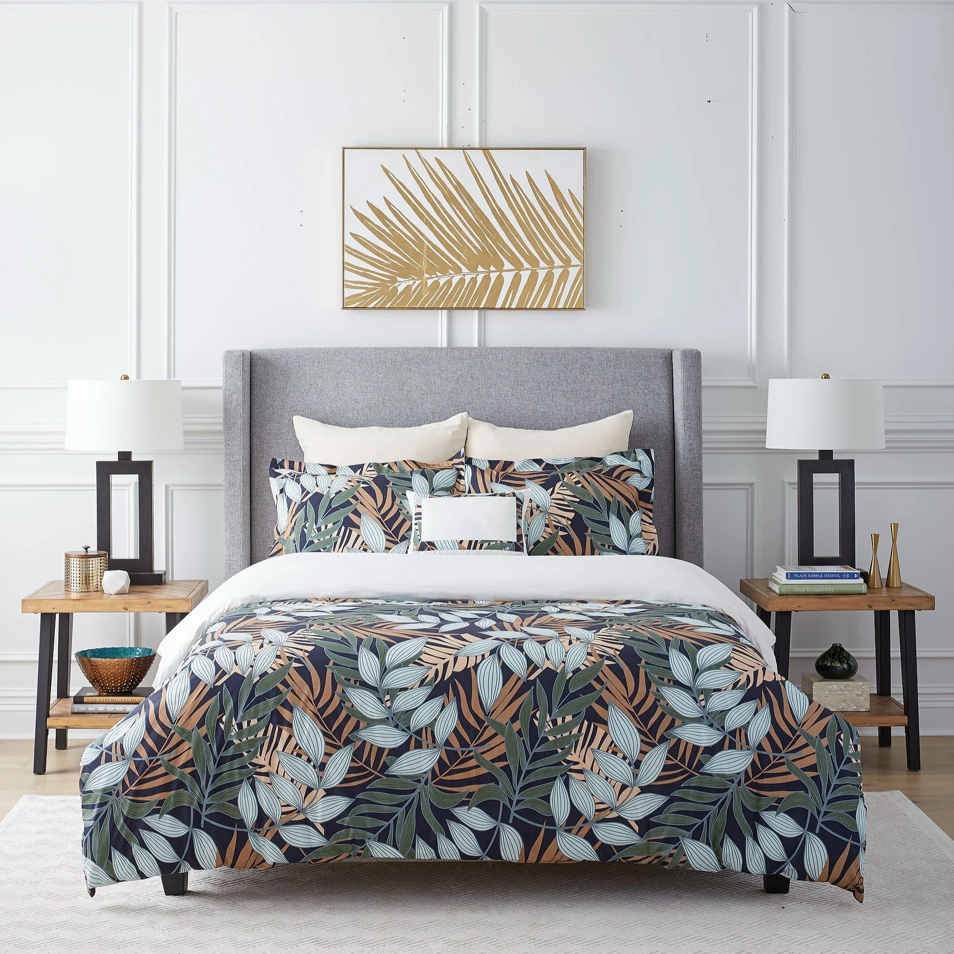 A neatly made bed with a gray upholstered headboard, patterned pillows, and a floral bedspread in front of white paneled walls with a gold-framed leaf artwork above. Two matching wooden nightstands with lamps and decorative objects flank the bed.