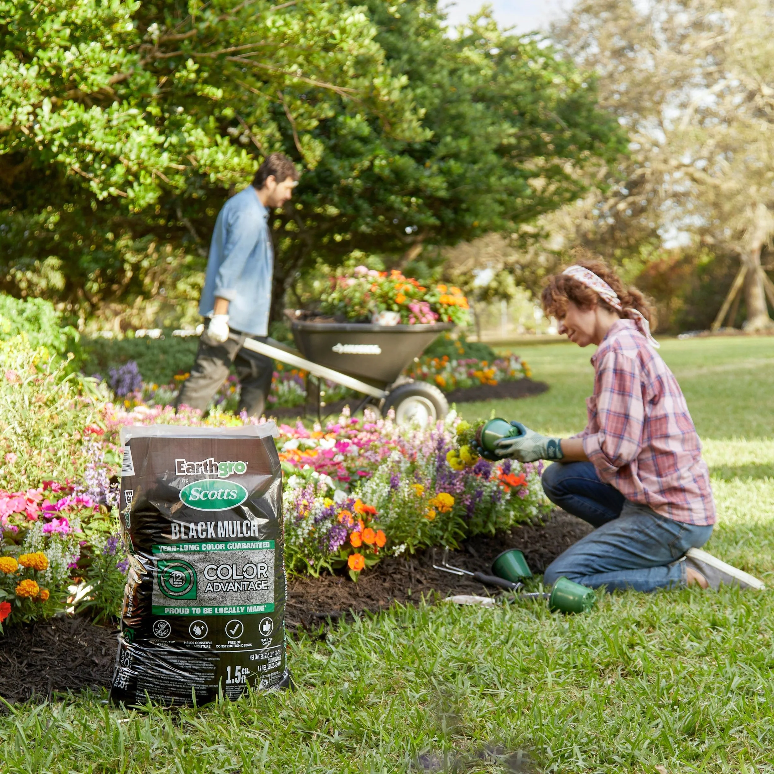 A woman kneeling on the grass planting flowers, with gardening tools and a bag of mulch nearby; a man in the background pushing a wheelbarrow filled with colorful flowers in a garden.