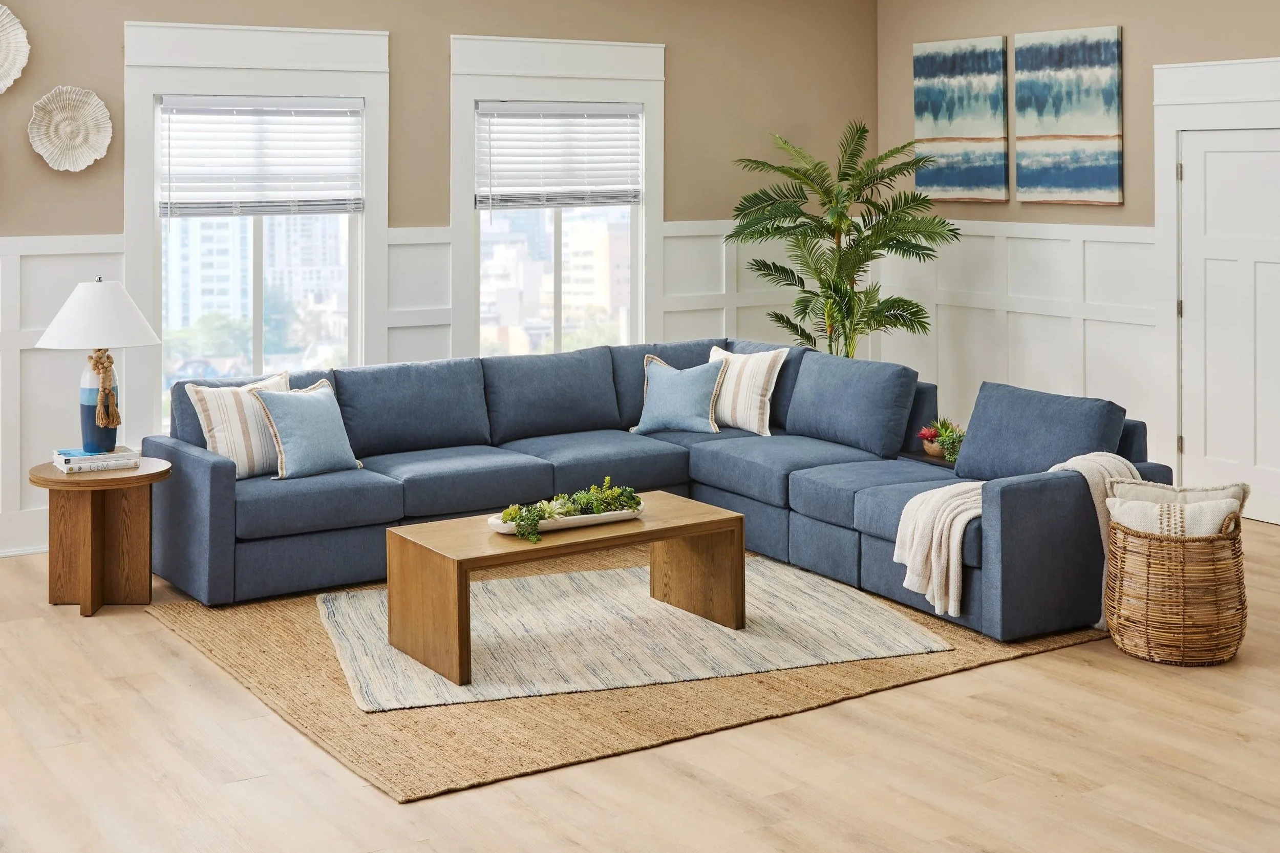 Living room with a blue sectional sofa, wooden coffee table, and side table with a lamp, beige walls with white trim, large windows with blinds, a green potted plant, and wall art featuring blue watercolor patterns.