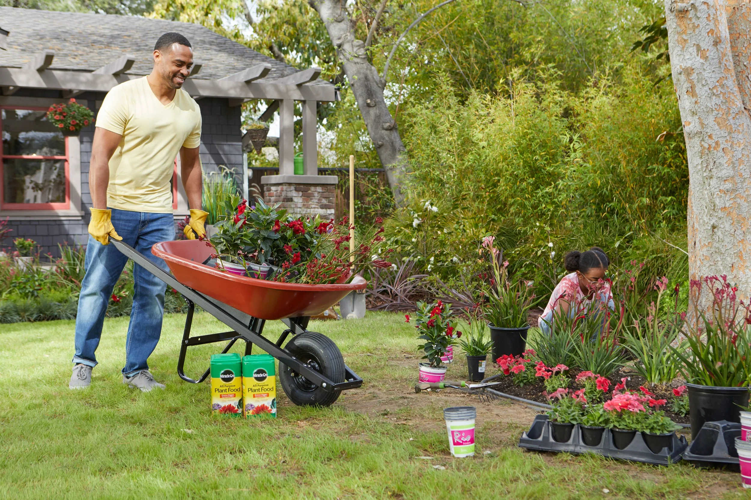 Two people planting flowers in a garden. One man with a yellow shirt and yellow gardening gloves pushing a wheelbarrow filled with plants. A woman kneeling on the ground, tending to potted flowers and plants. Several plant containers and gardening su