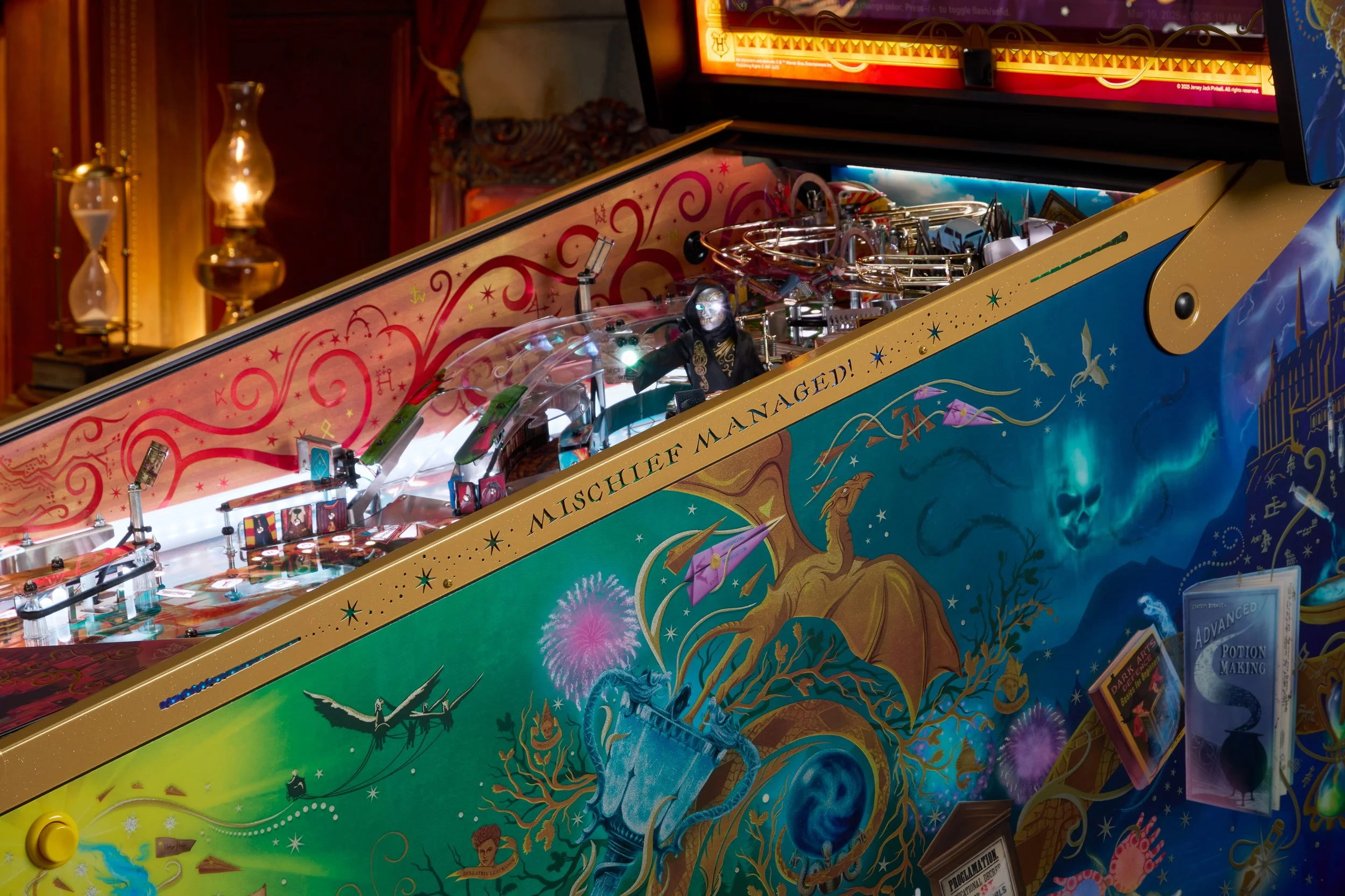 Close-up of a colorful pinball machine with the words 'Mischief Managed' on the side, featuring artwork of a dragon, a phoenix, and magical themes, with some flippers and bumpers visible.