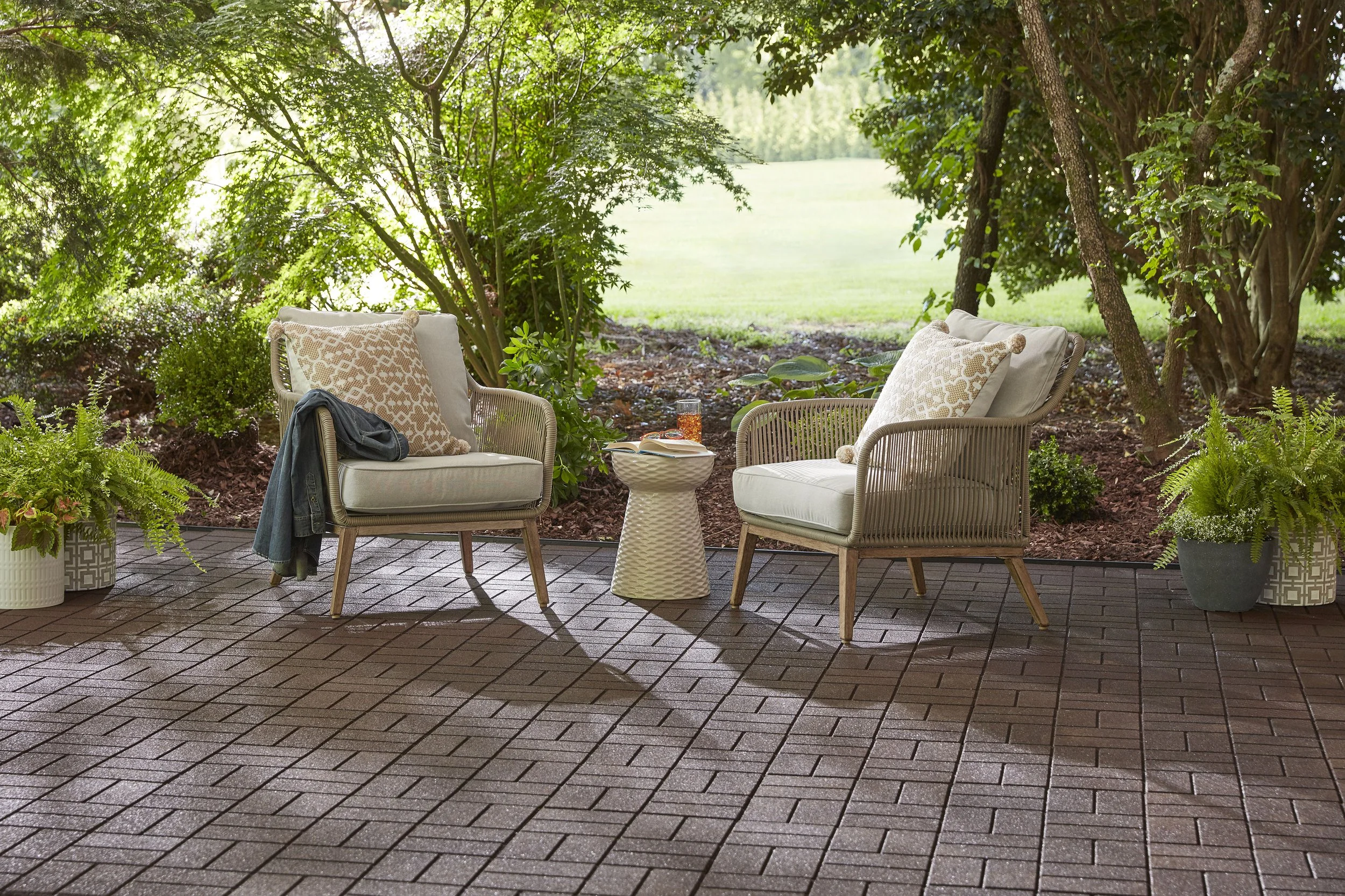 Two beige outdoor chairs with pillows, a small white side table with a book and a glass of drink, set on a brick patio surrounded by green plants and trees, overlooking a grassy yard.