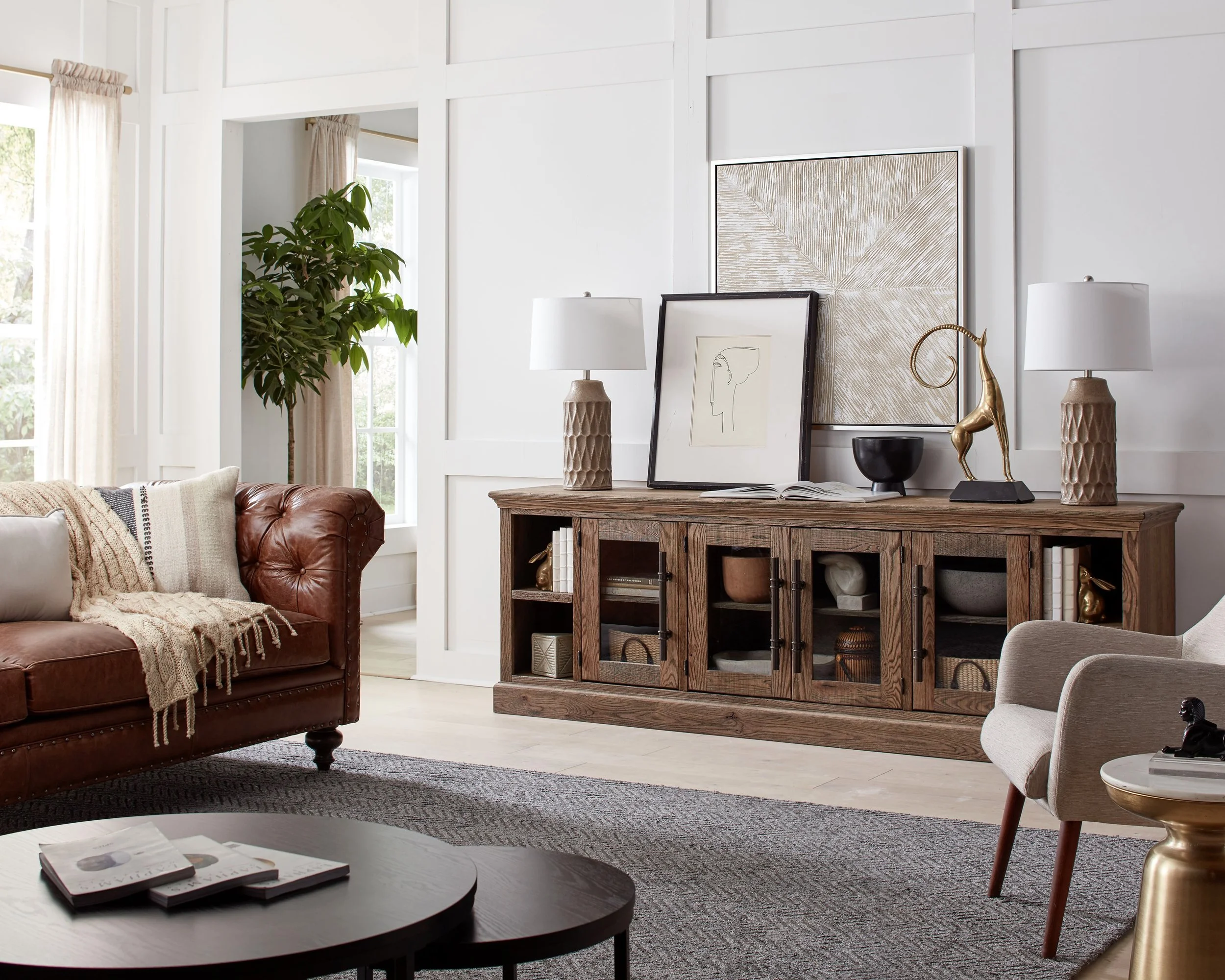 A cozy living room with a leather sofa, a wooden sideboard decorated with lamps, artwork, and decorative items, a round coffee table, an armchair, a striped throw blanket, a rug, and large windows with light curtains, and a tall plant in the corner.