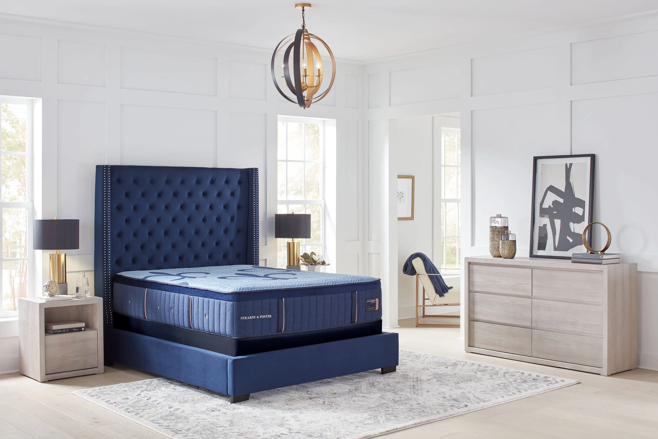 A modern bedroom with a blue upholstered bed, bedside tables with black lamps, a dresser with decorative items, and large windows with natural light.