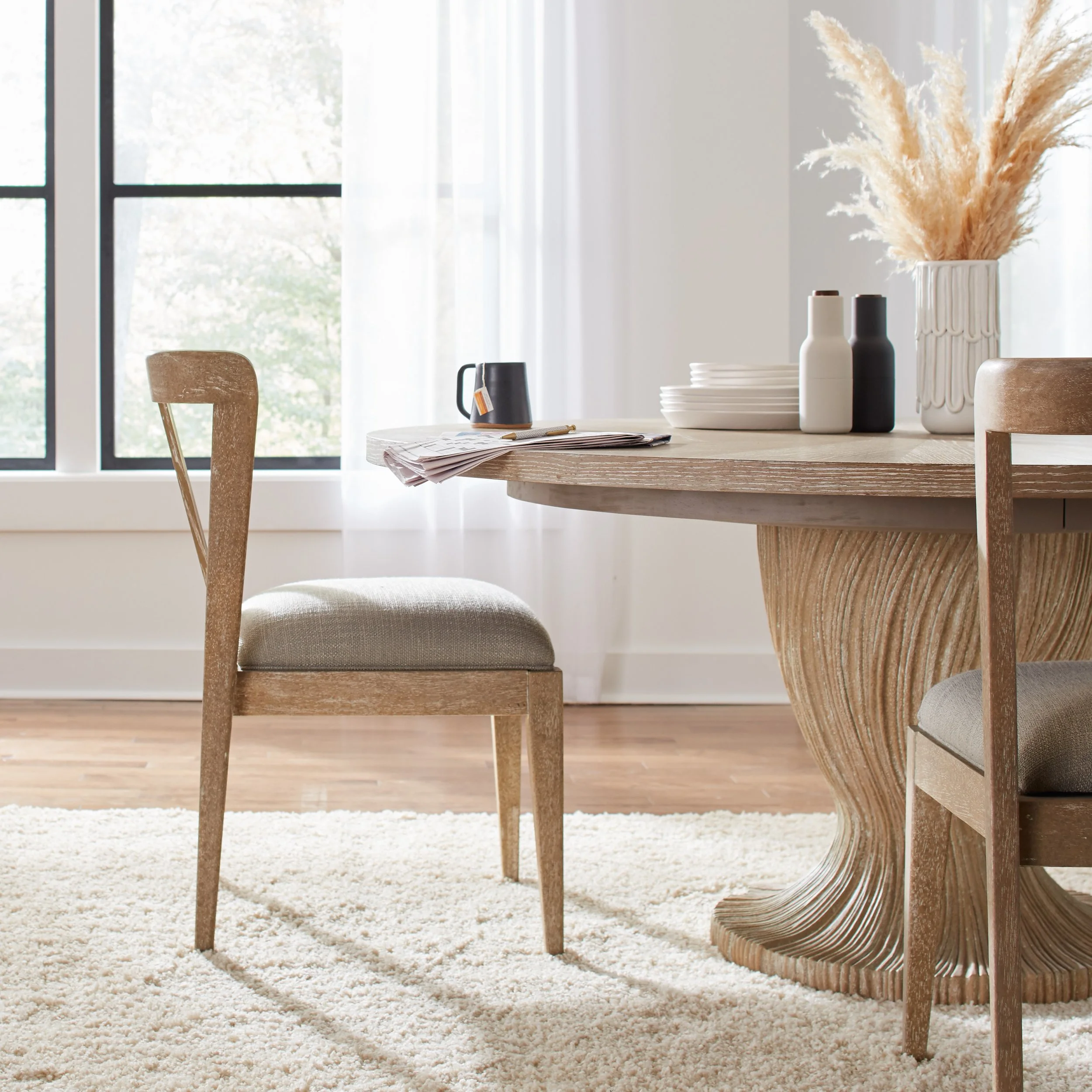 A dining room with a round wooden table, beige upholstered chairs, a white shag rug, and a large window with sheer curtains. The table has a vase with pampas grass, plates, and bottles.