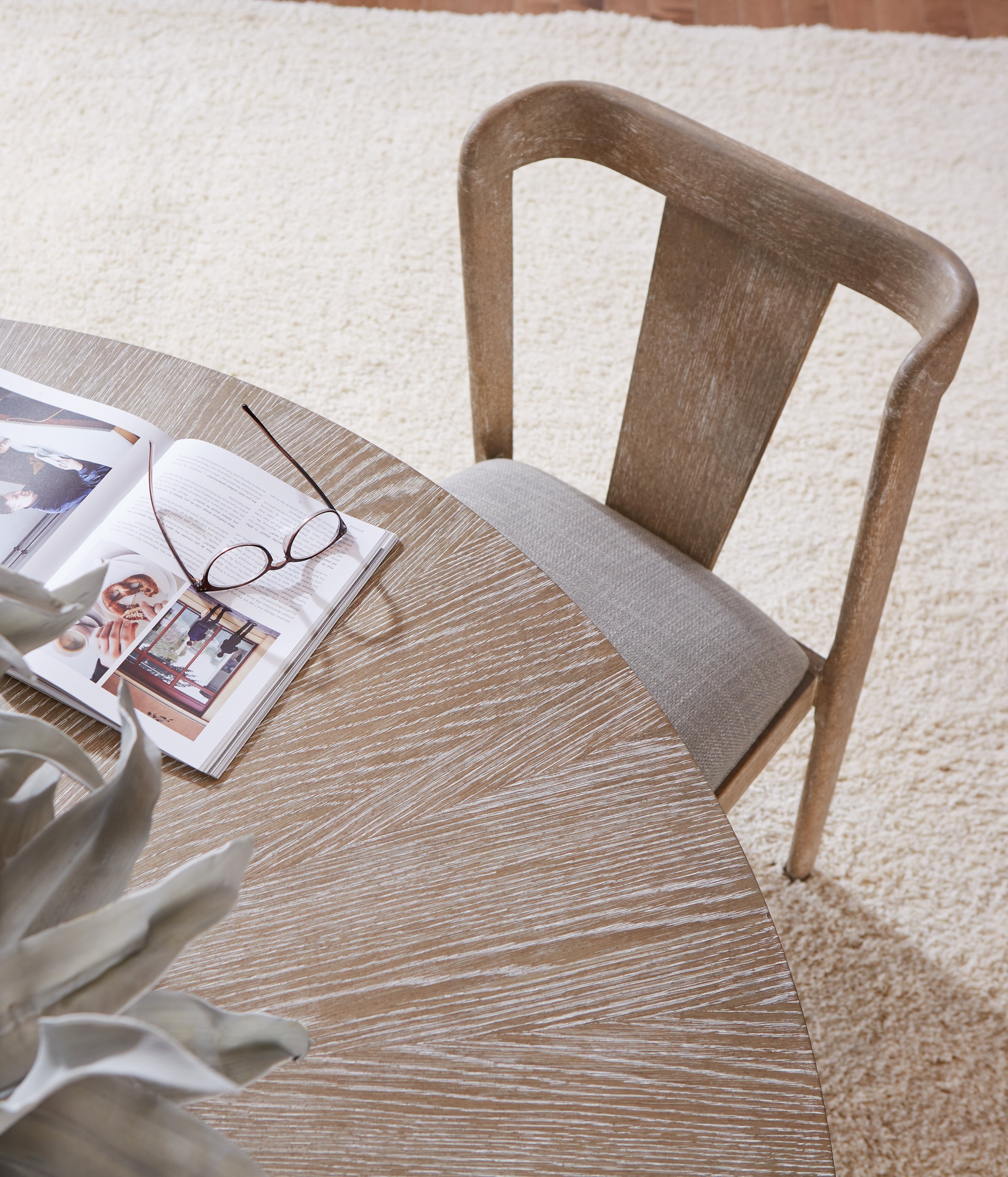 A wooden chair with a gray cushion placed next to a round wooden table. On the table, there is an open magazine and a pair of glasses. A potted plant with large leaves is partially visible in the foreground. The setting appears to be indoors on a bei