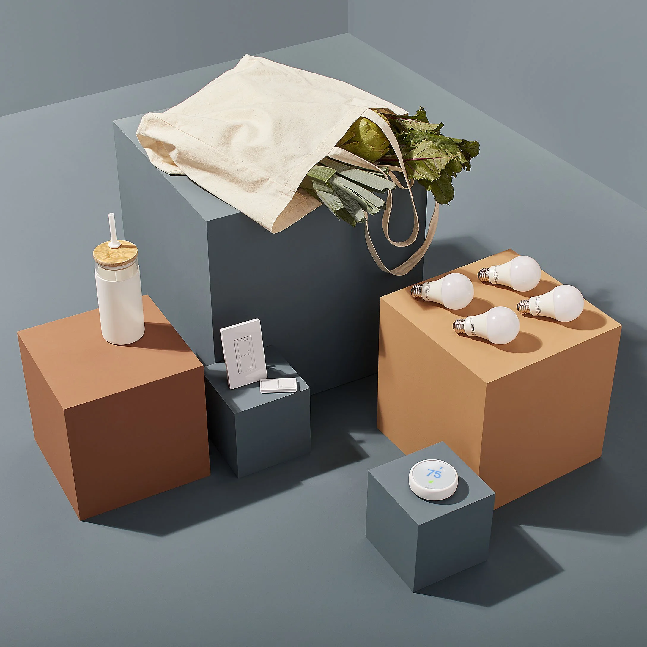 A collection of smart home devices and accessories on geometric pedestals, including a reusable straw, a tote bag with vegetables, four light bulbs, a smart thermostat in a white circular device displaying 75°, and a relay switch.