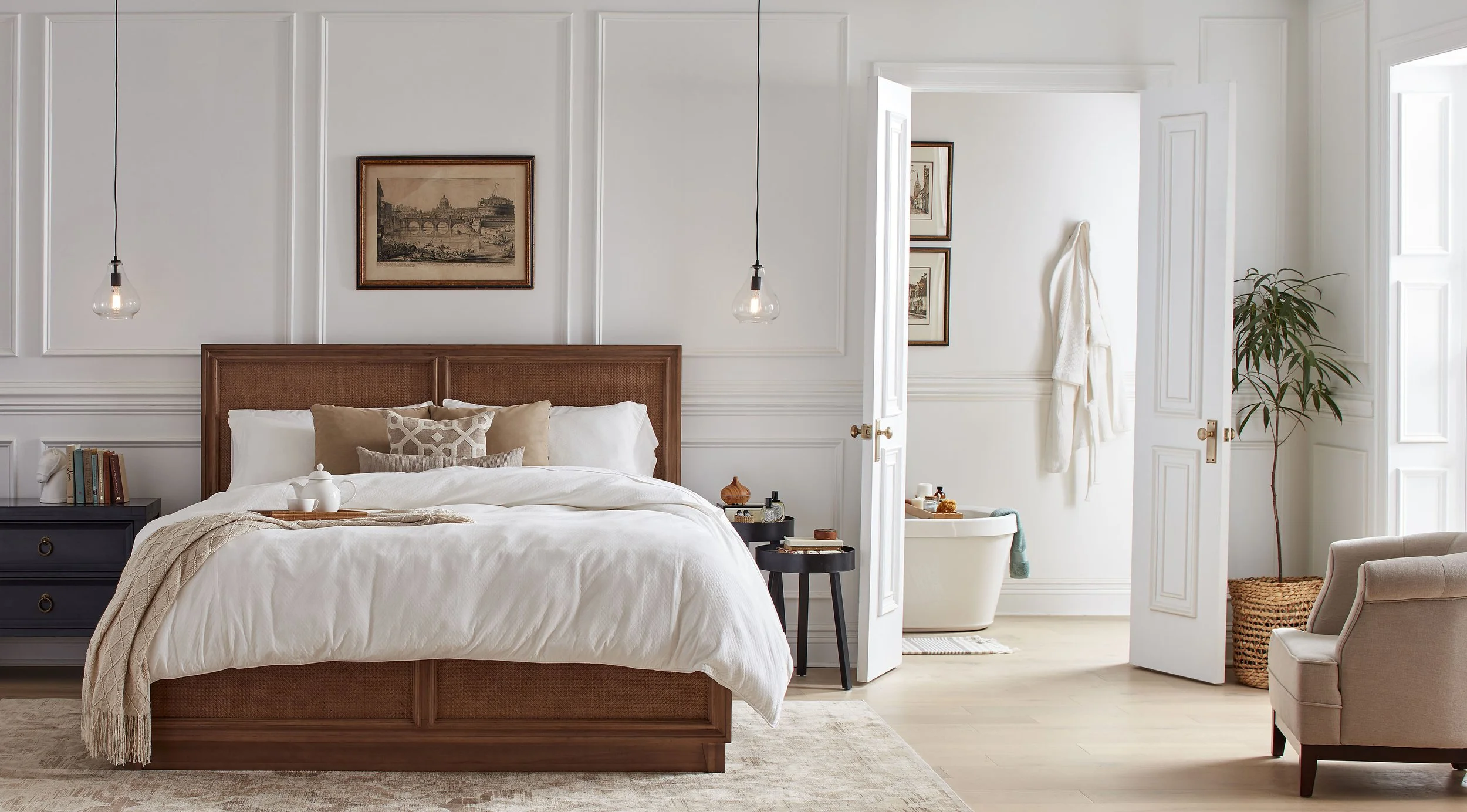 A bedroom with a large bed featuring white and beige bedding, a wooden headboard, and two hanging pendant lights. There are side tables with decor, a framed picture above the bed, and a sitting area with an armchair. An open door reveals a bathroom w