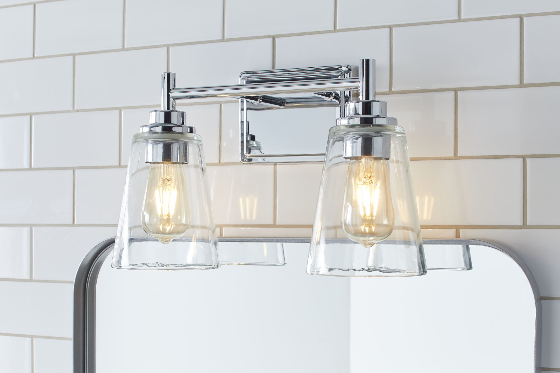 Two exposed lightbulbs hanging from a chrome fixture above a mirror with a white tiled wall in the background.