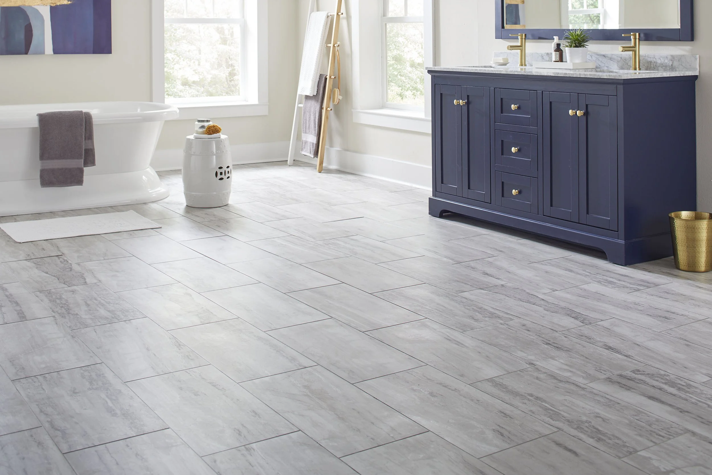 A modern bathroom with white walls and large windows letting in natural light. There is a white bathtub with a gray towel draped over the side. A white ceramic stool with spa items is beside the tub. A wooden towel ladder with towels is near the wind