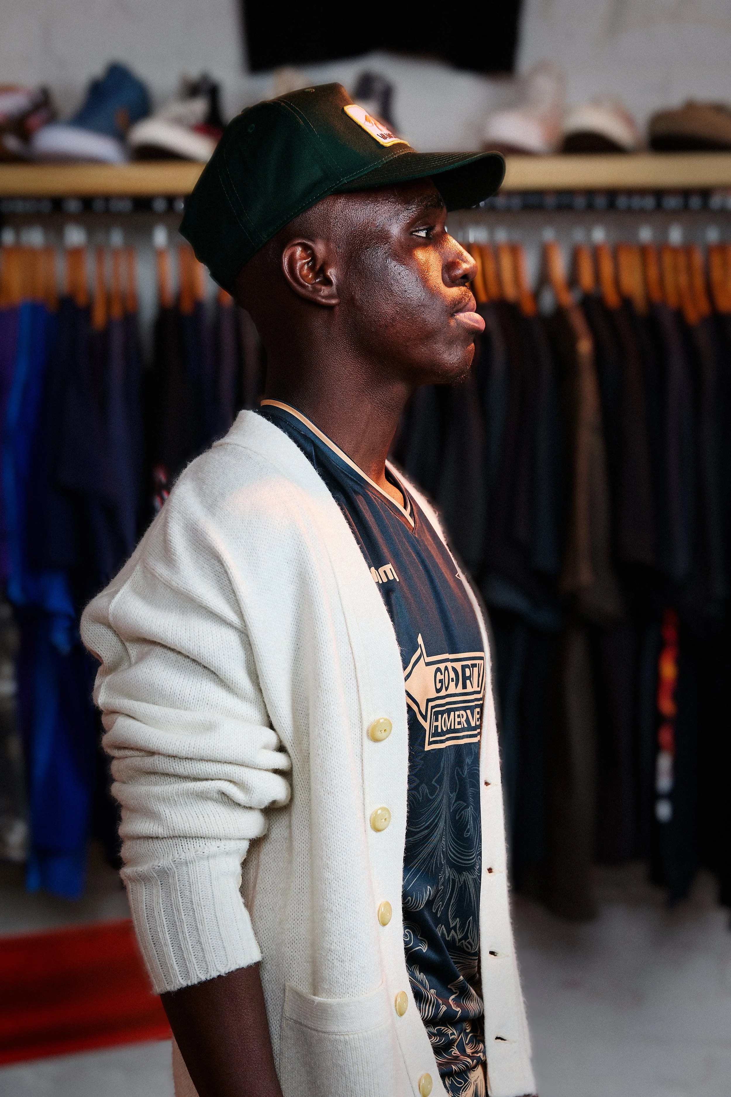 A man in a black cap, white cardigan, and sports jersey standing inside a store with clothes hanging behind him.