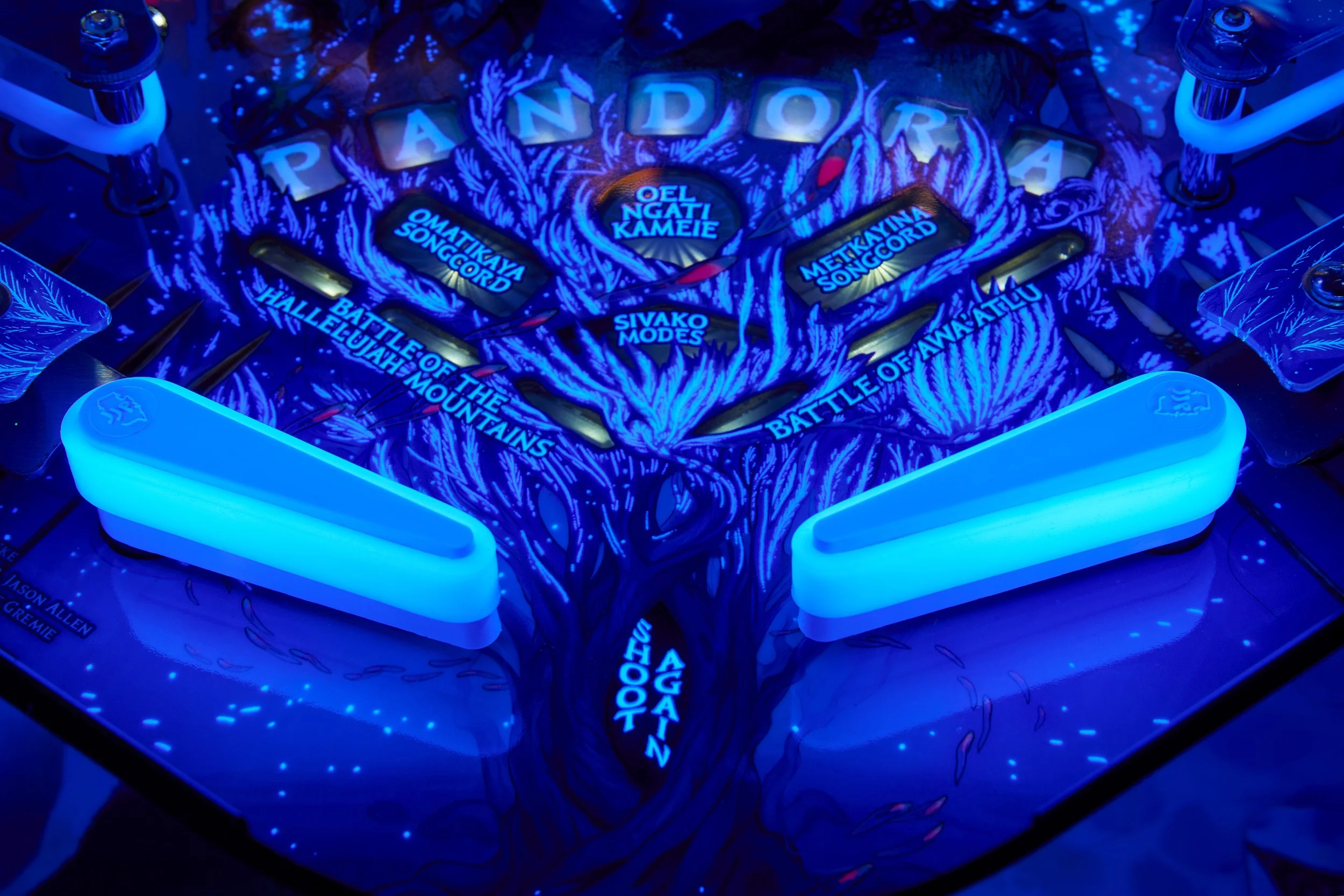 A close-up of a pinball machine with a blue-lit theme featuring a tree design, battle of the mountains, and song records, with parts of the pinball table's bumpers and flippers shown.