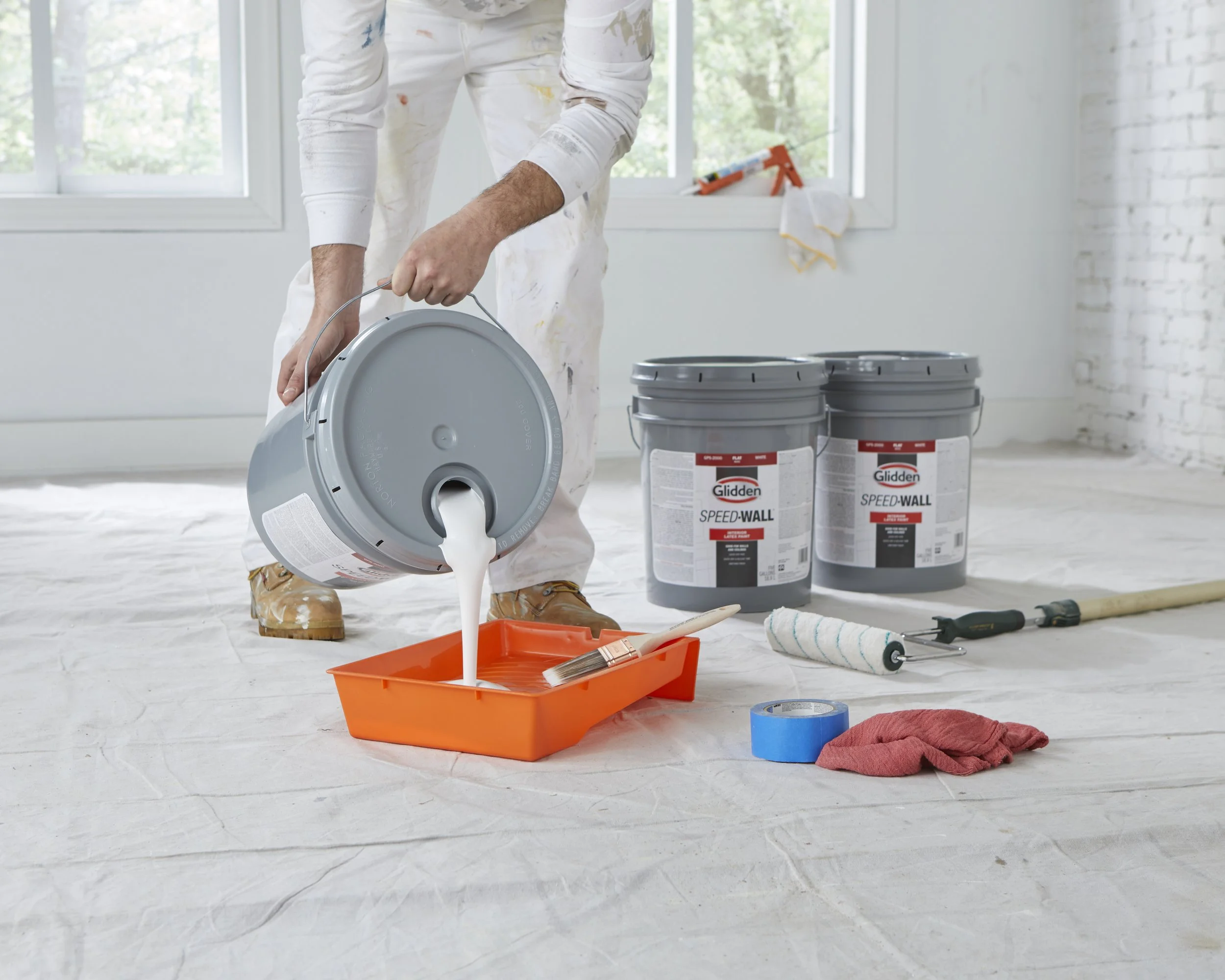 A person painting inside a room with a bucket of white paint, paintbrush, taped plastic sheet on the floor, and painting supplies nearby.