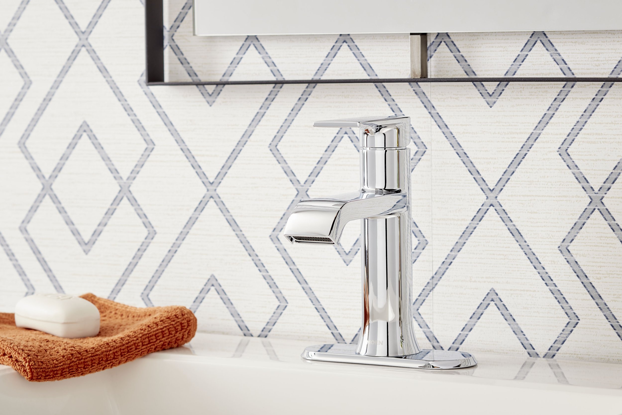 Close-up of a modern chrome faucet on a bathroom sink, with a bar of soap on an orange towel and a mirror above, set against a white wall with a diamond-patterned tile backsplash.