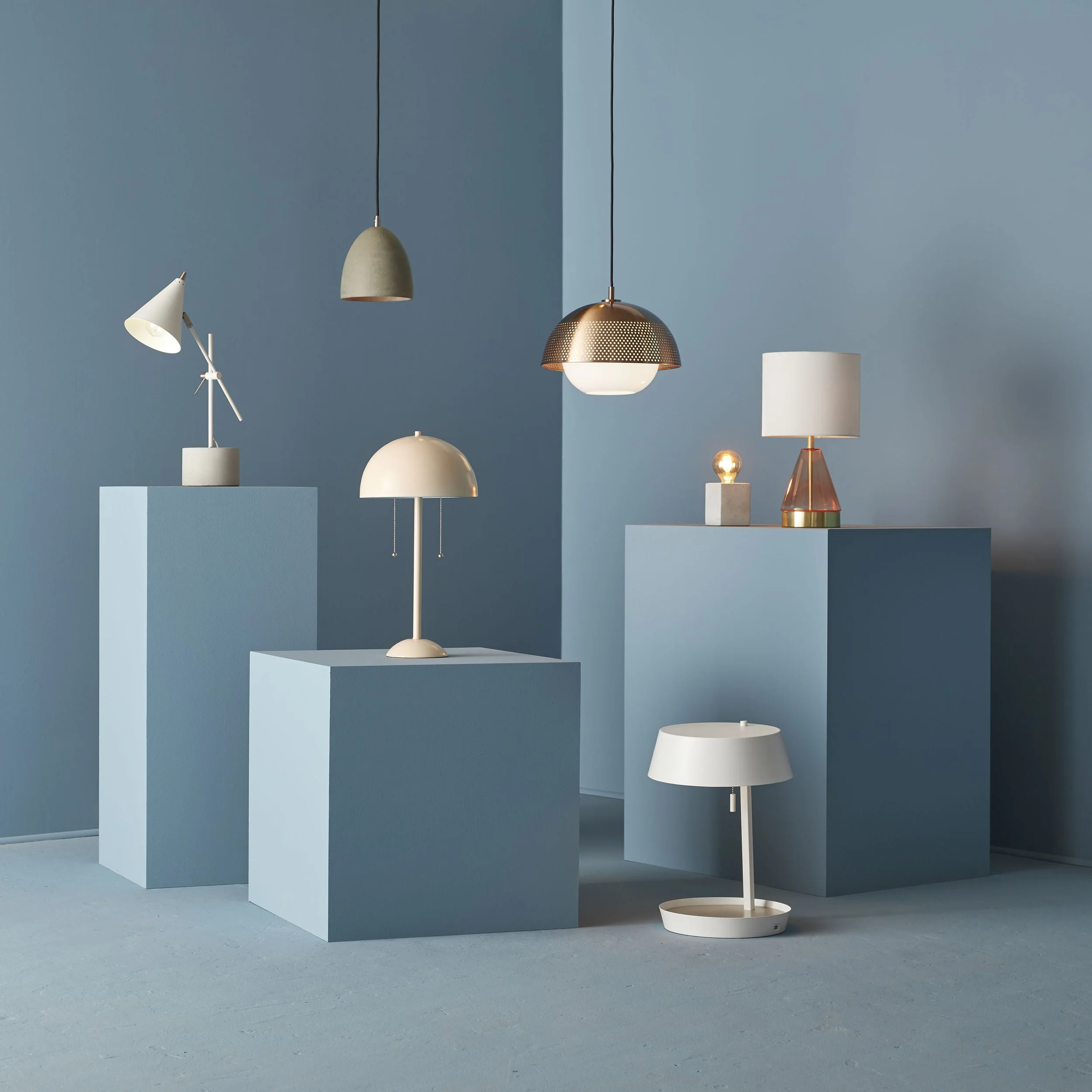 Display of various modern lamps on blue pedestals and against a blue wall, with contrasting hanging and table lamps in a minimalist arrangement.