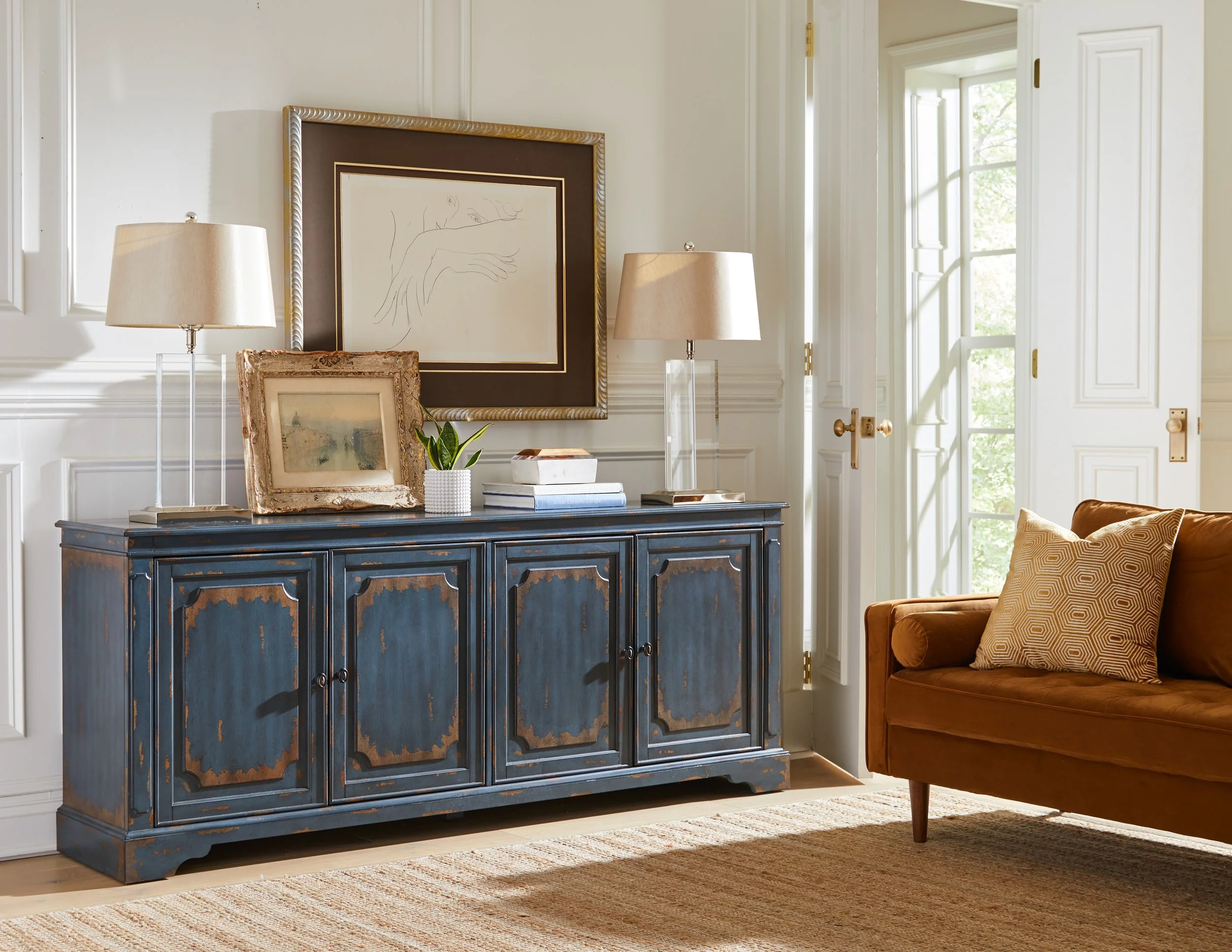 A cozy living room corner with a distressed blue sideboard, artwork, lamps, a potted plant, and a brown sofa with patterned cushions, next to a large window.