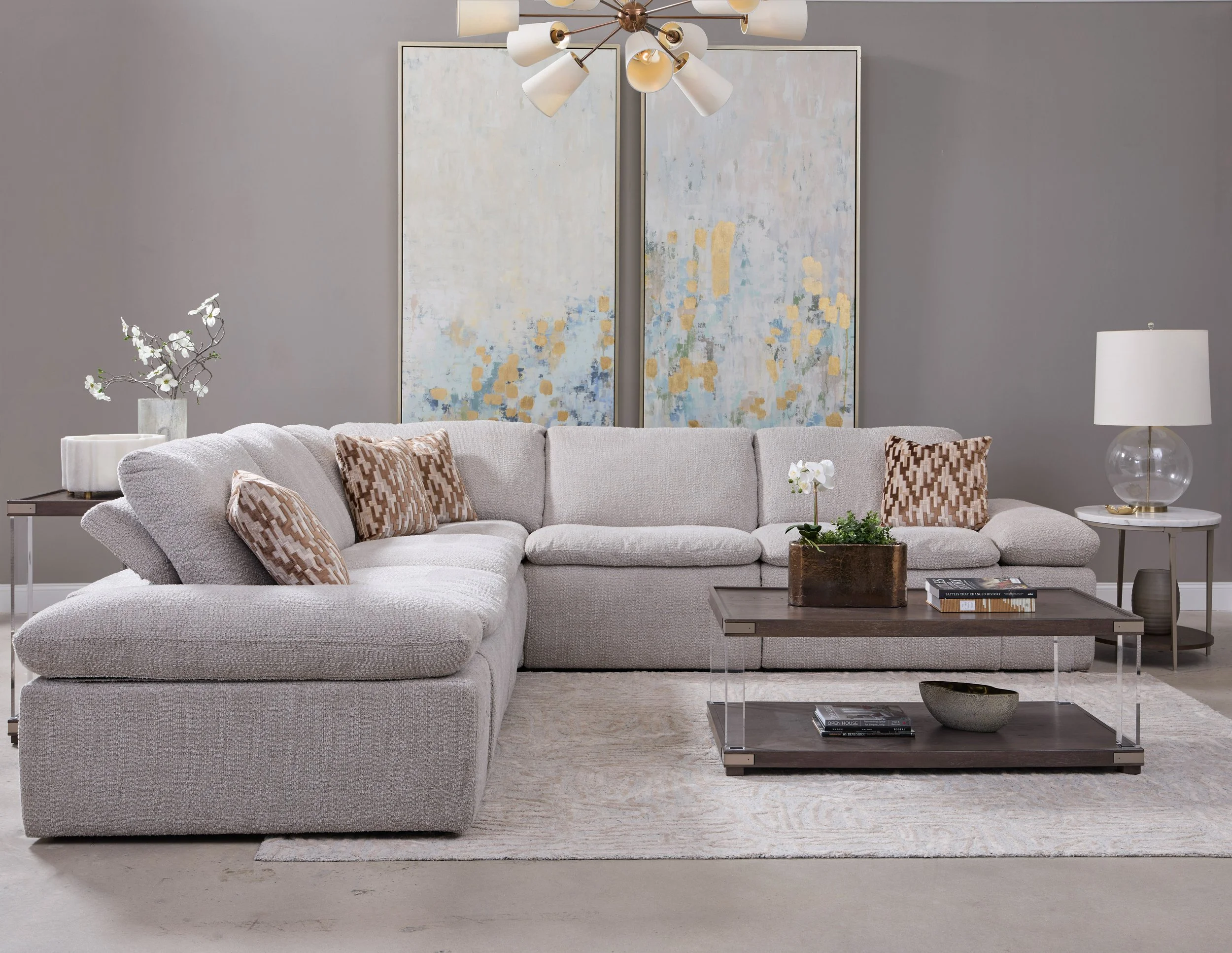 Living room with a large white sectional sofa, decorative pillows, a dark brown coffee table with a potted orchid, side tables with lamps and decorative objects, and an abstract painting on a gray wall.