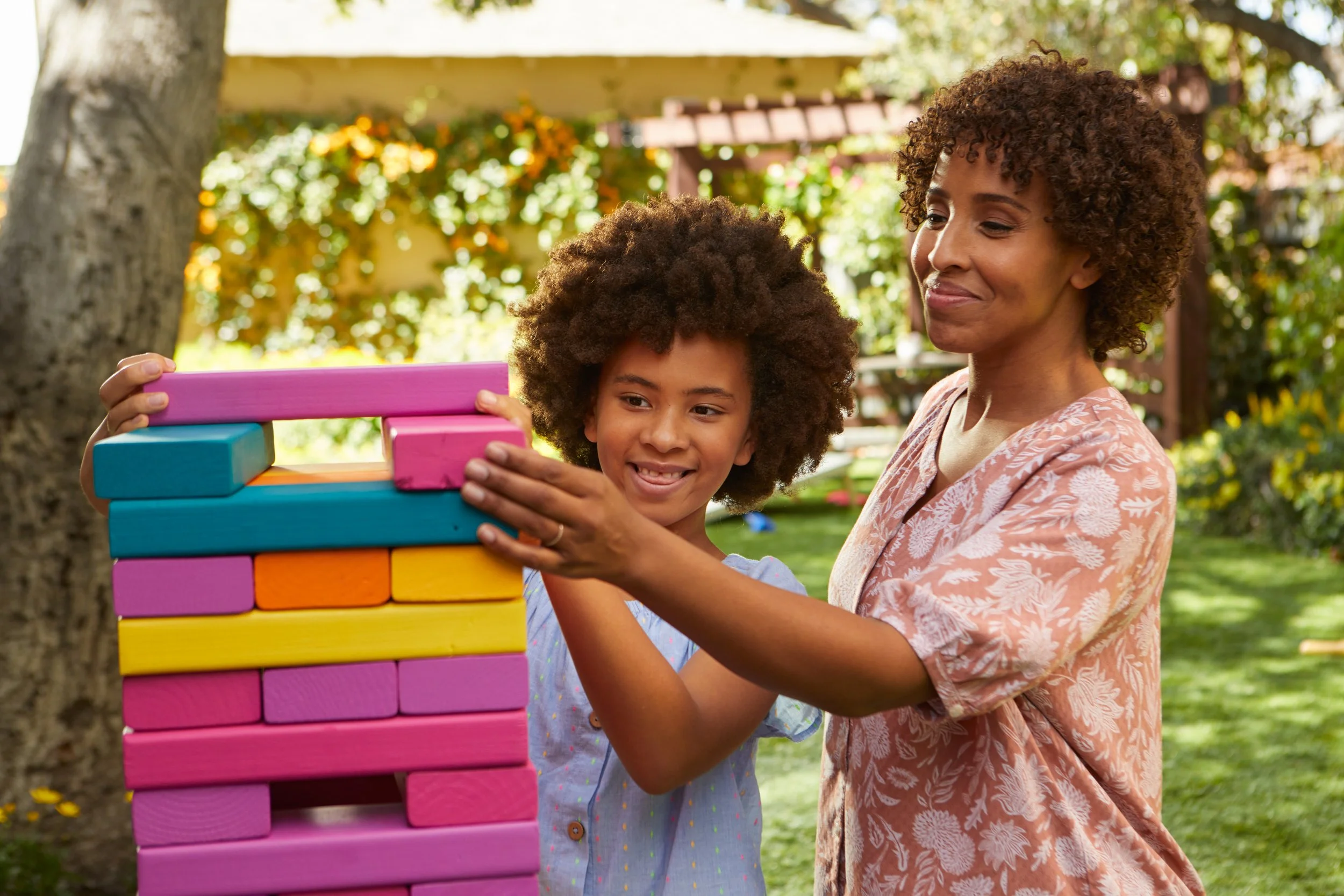 A woman and a girl with curly hair playing giant Jenga outdoors in a garden with trees and flowers.