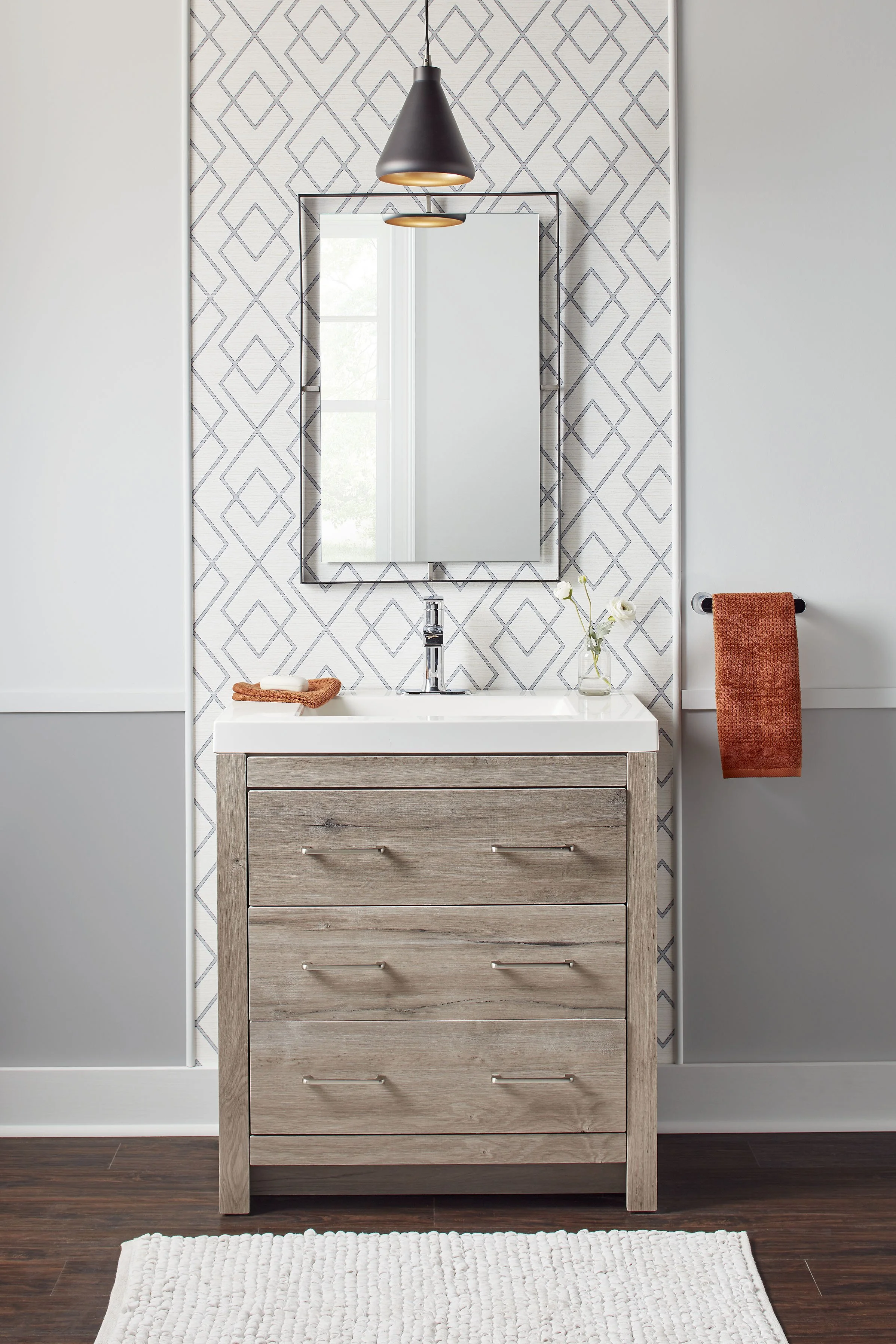 A modern bathroom vanity with a wood finish, featuring four drawers, a white countertop with a built-in sink, a mirror, a black pendant light, a small flower vase, and a towel hanging on a black bar, with a geometric patterned wall behind.