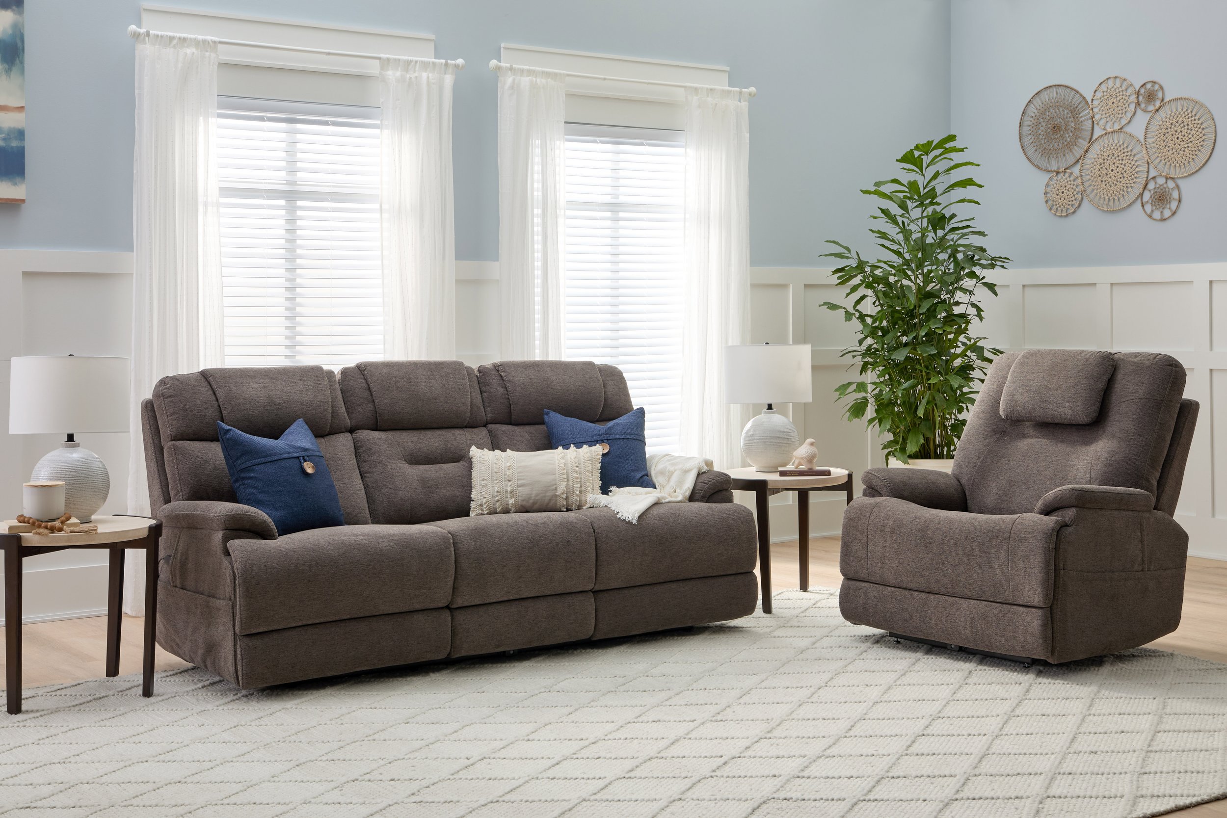 Living room with gray sofa and recliner, white curtains, green plant, decorative wall art, two white table lamps, beige rug.