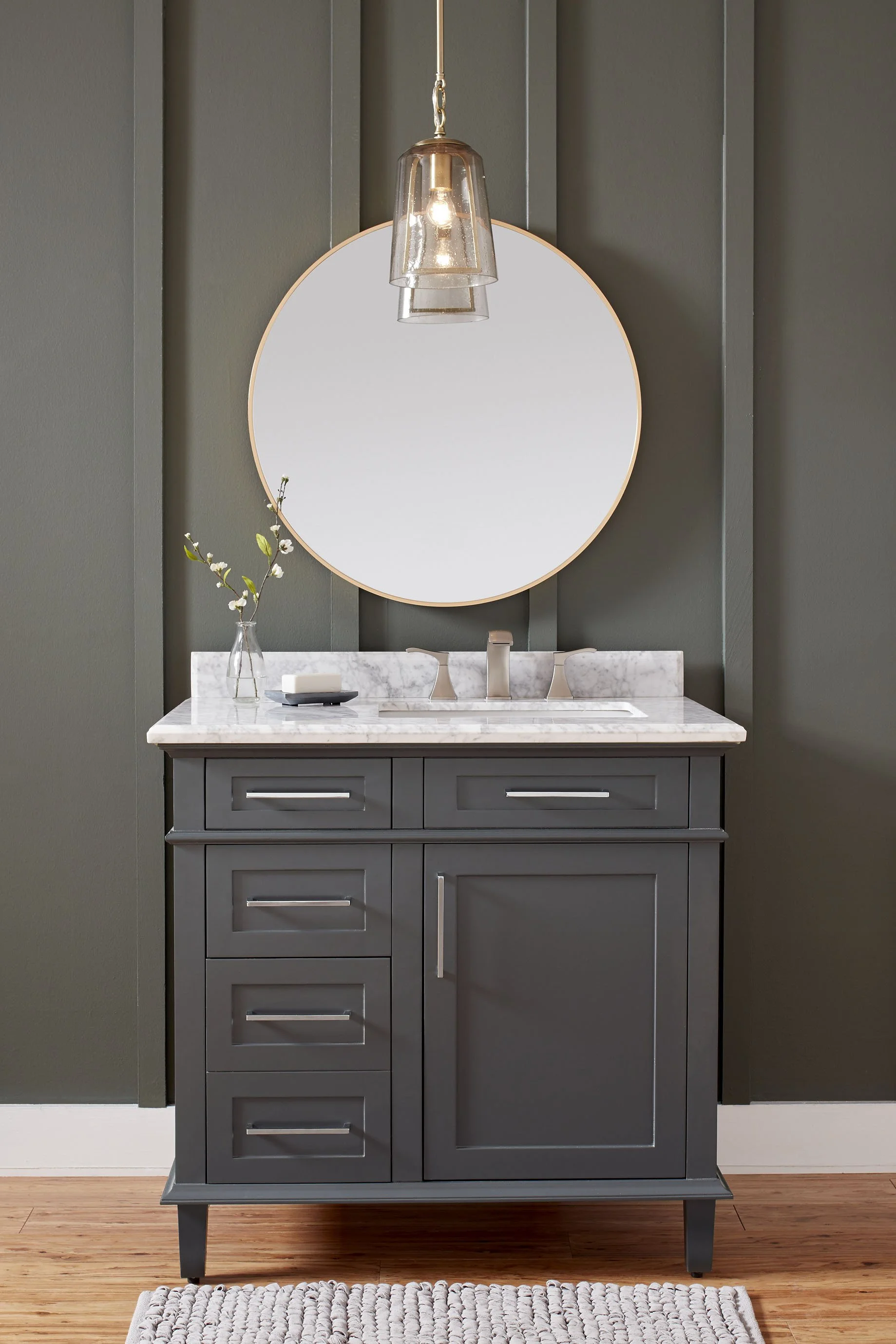 A bathroom vanity with gray cabinet, a white marble countertop, a round mirror above, a glass pendant light, a small vase with a sprig, and a bar of soap.