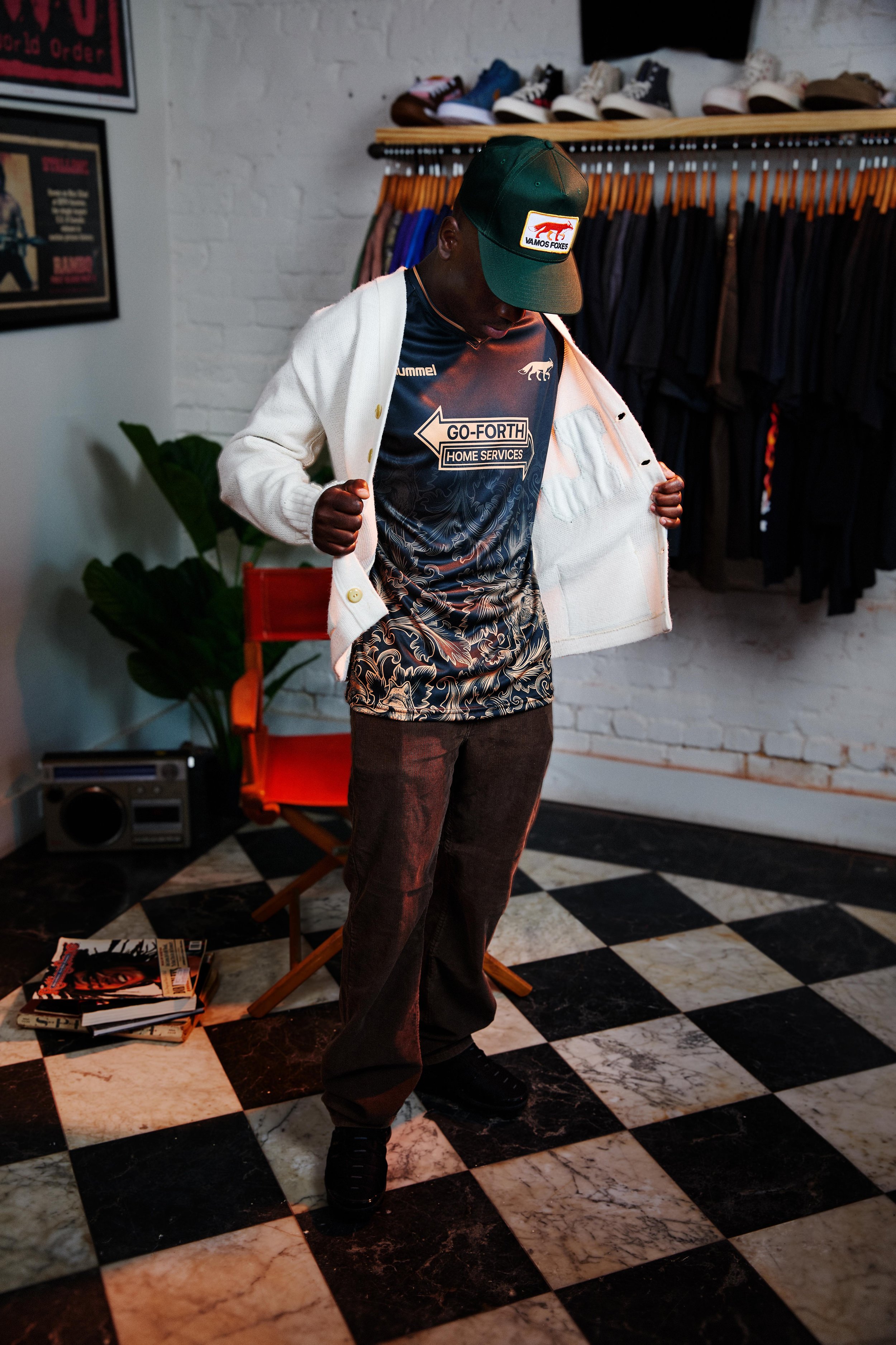 A person wearing a baseball cap, a patterned shirt, and a white jacket, standing in a room with checkered floors, a coat rack, a chair, and a small collection of magazines.