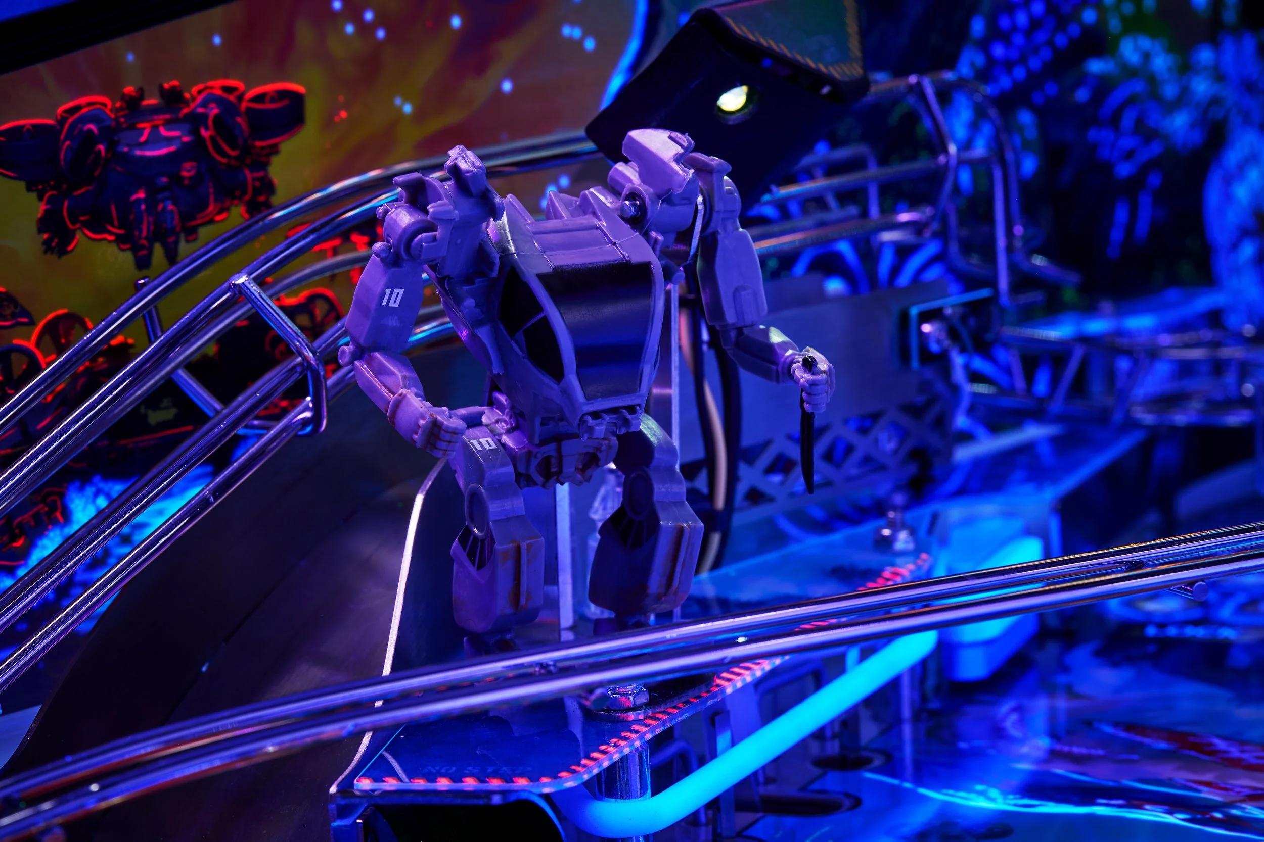 Close-up of a pinball machine featuring a robotic humanoid figure on the playfield, illuminated by blue and purple lights, with other gaming artwork in the background.
