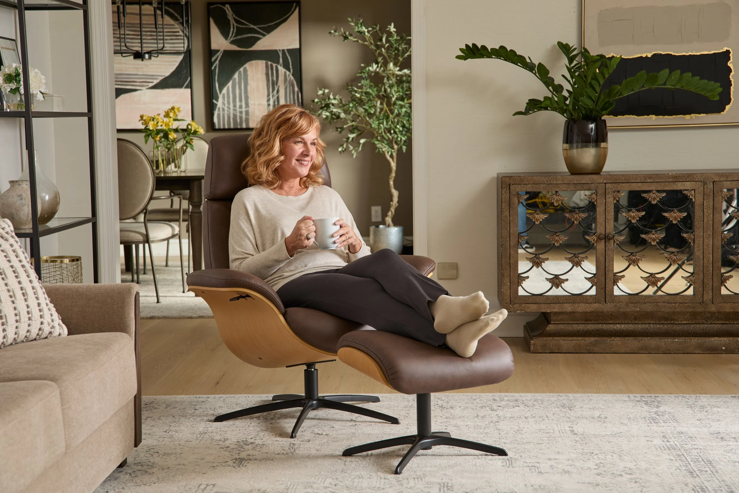 A woman with curly blonde hair sitting in a modern living room on a brown lounge chair with an ottoman, holding a white mug, smiling. The room features neutral tones, a beige sofa with a patterned pillow, a decorative console with black doors, a larg