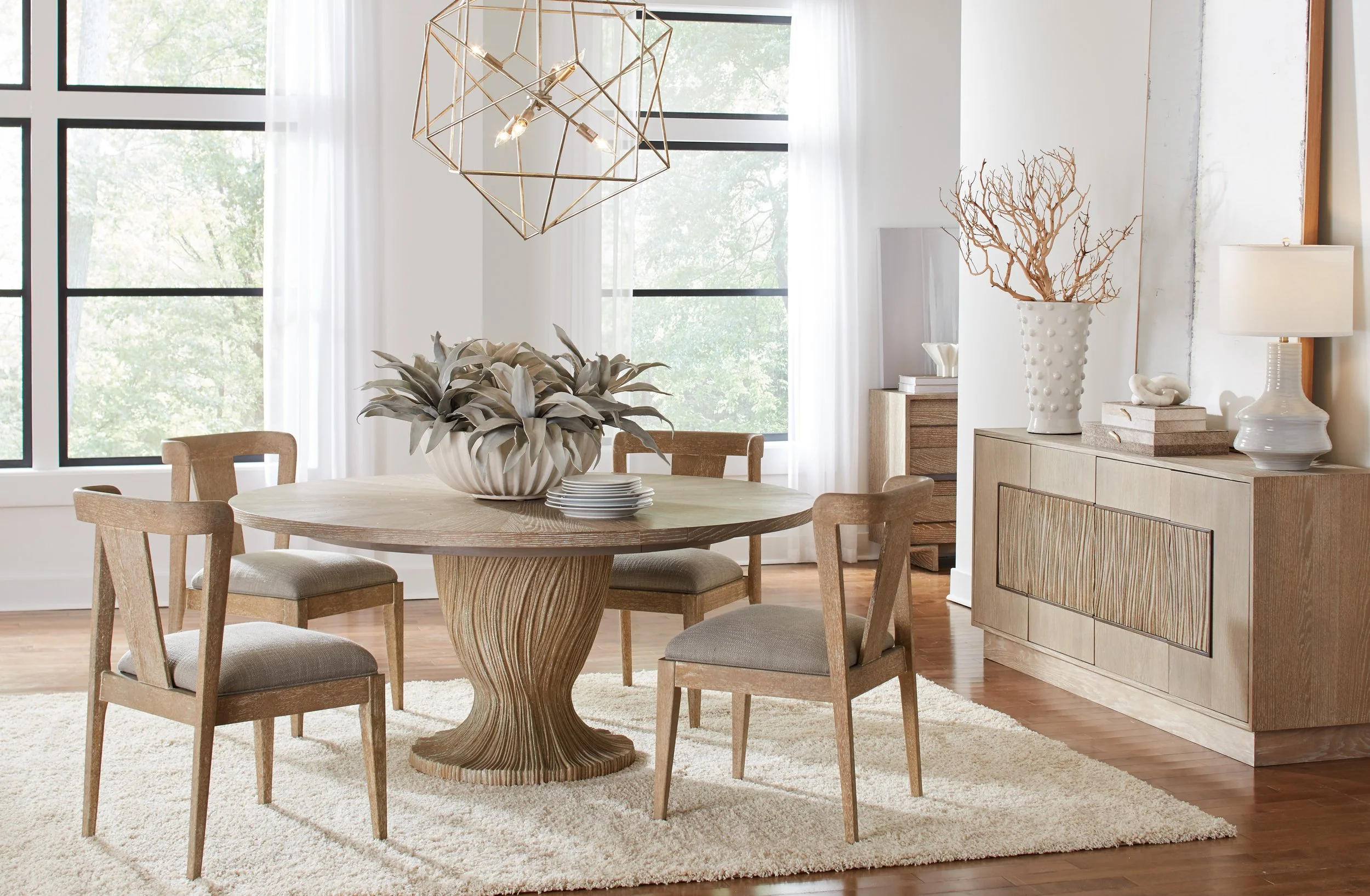 Dining room with a round wooden table, four matching chairs, a large decorative vase with foliage, and a sideboard with decorative items, illuminated by natural light from large windows.