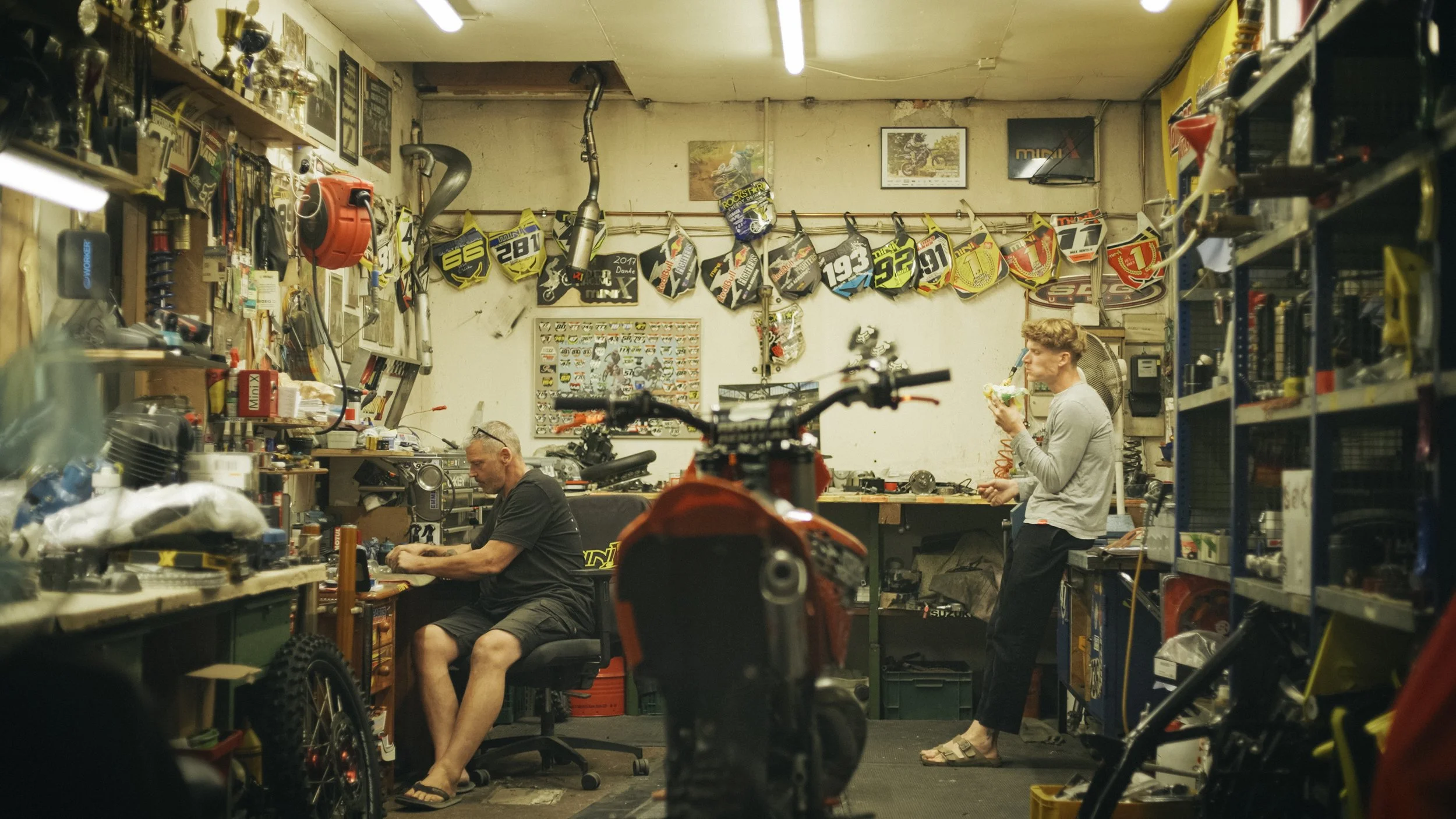 Werkstatt mit zwei Männern, einem sitzt an einem Schreibtisch und arbeitet, der andere steht und isst. Motorradurausgestattet mit Helmen und Teilen hängen an der Wand, Regale voller Werkzeuge und Zubehör sind sichtbar.