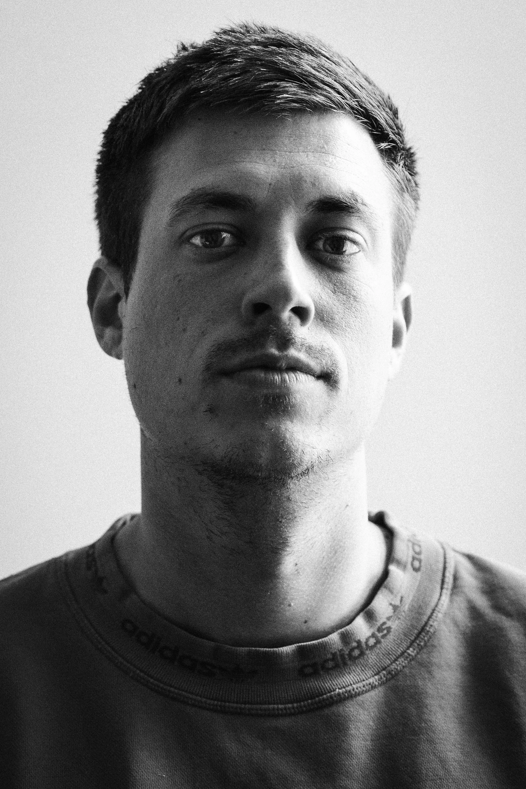 Robin Fischer Portrait, Schwarz-weiß-Porträt eines jungen Mannes mit kurzen Haaren, einige Gesichtshaare, trägt ein Adidas-T-Shirt.