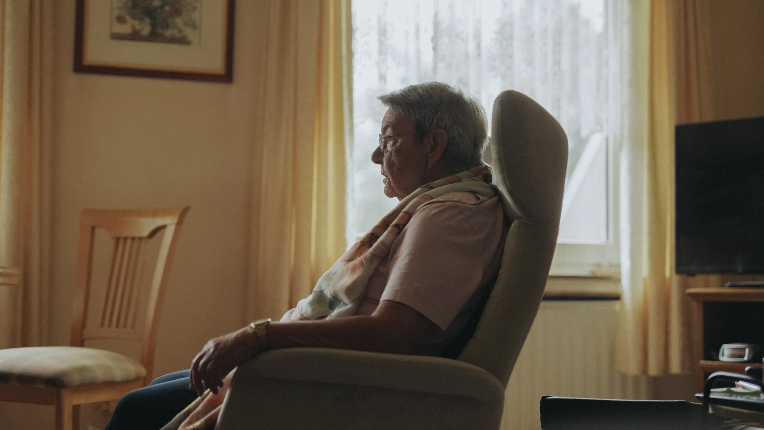 Älterer Mann sitzt nachdenklich in einem Sessel vor einem Fenster mit durchsichtigen Vorhängen in einem Wohnzimmer, seitlich betrachtet.
