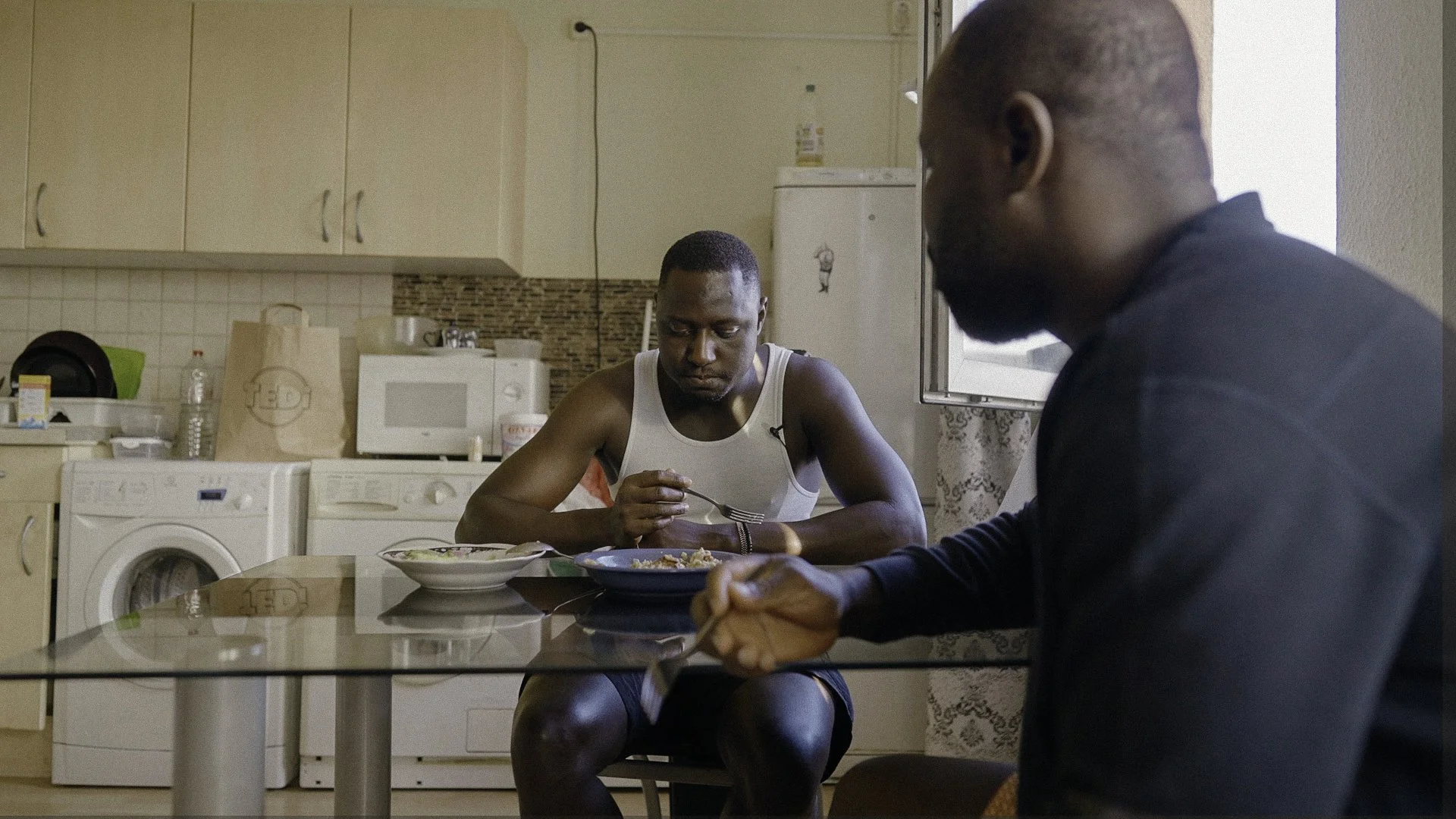 Zwei Männer sitzen während des Essens in einer Küche. Einer trägt ein weißes Tanktop, der andere ein schwarzes Hemd.