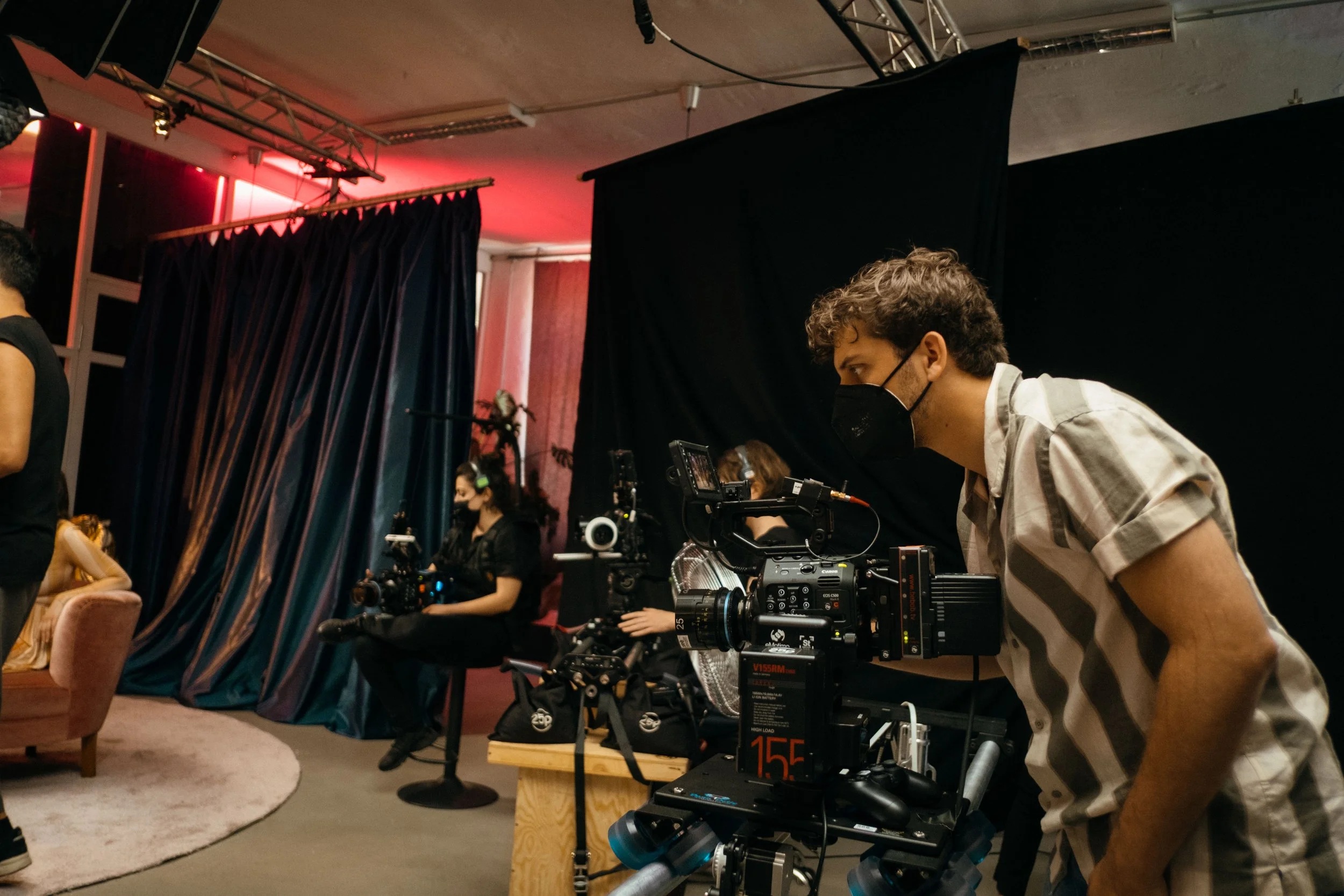 Ein Kinotechniker mit Maske arbeitet an einer Kinokamera in einem dunklen Raum, mit Vorhängen im Hintergrund, während andere Personen im Hintergrund das Filmset vorbereiten.