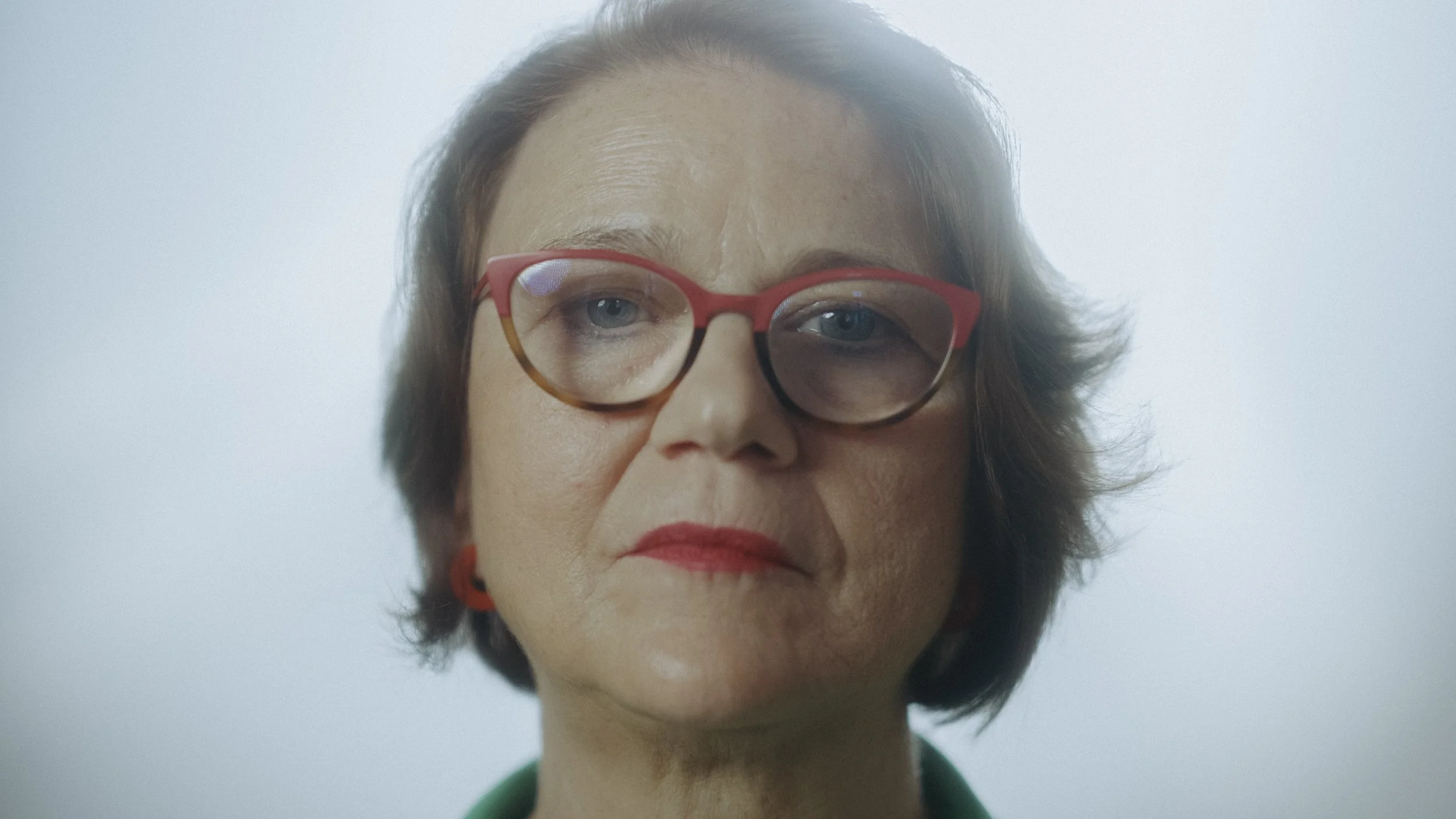 Nahaufnahme eines Erwachsenen mit Brille und roten Ohrringen. Hintergrund ist hell und unscharf.