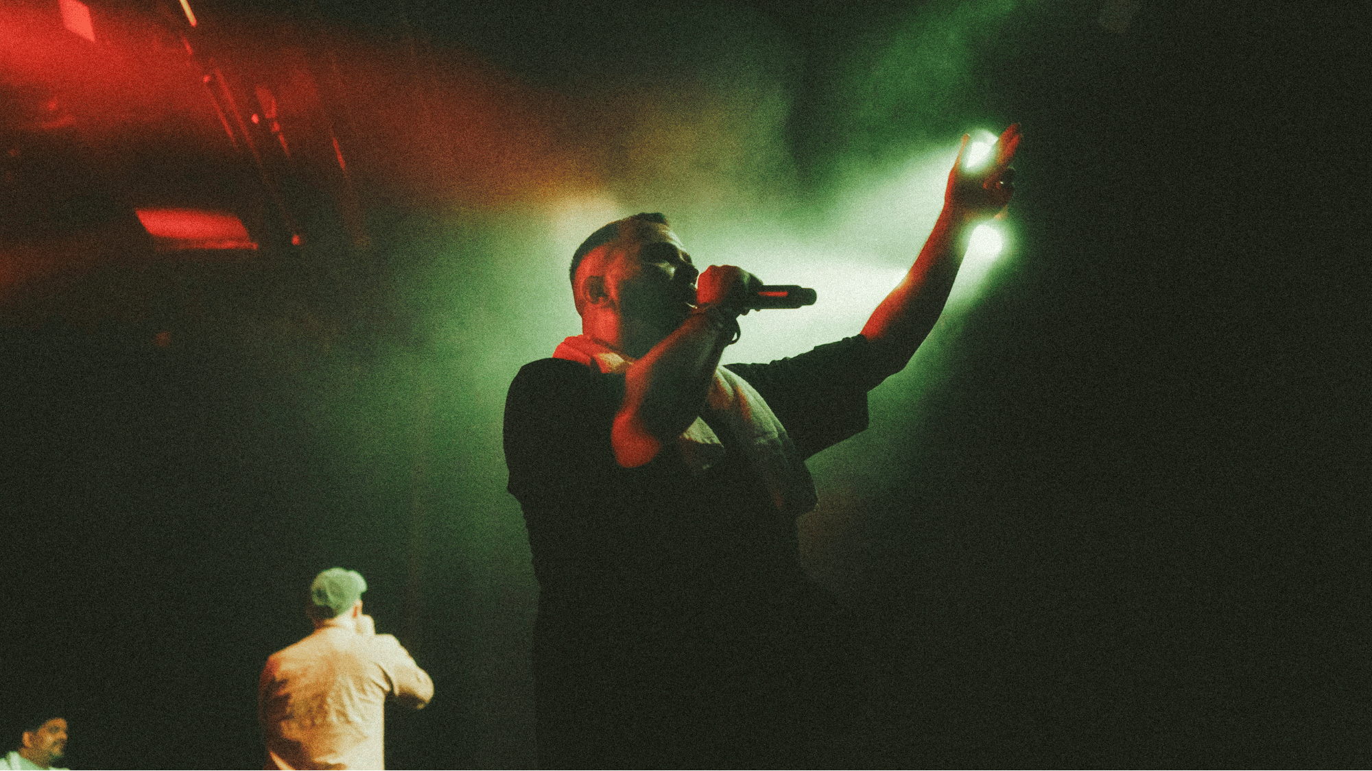 Ein Mann singt mit einem Mikrofon auf einer Bühne, während er in eine Hand in die Luft hebt. Es sind einige Menschen im Hintergrund, die auf der Bühne beobachten.