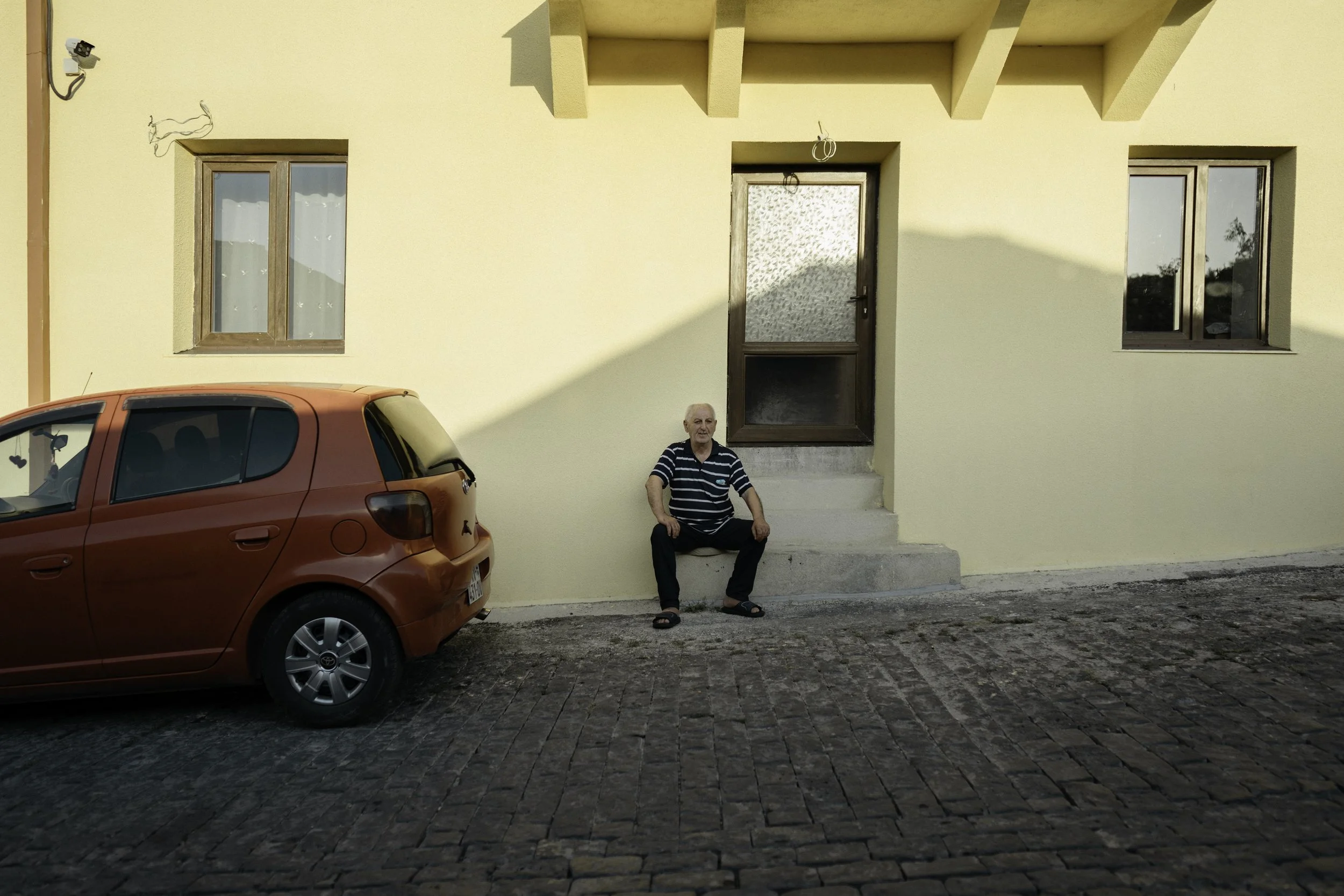 Ein älterer Mann sitzt auf den Stufen vor einem gelben Haus mit zwei Fenstern und einer Tür. Ein rotes Auto steht auf gepflastertem Boden neben ihm.