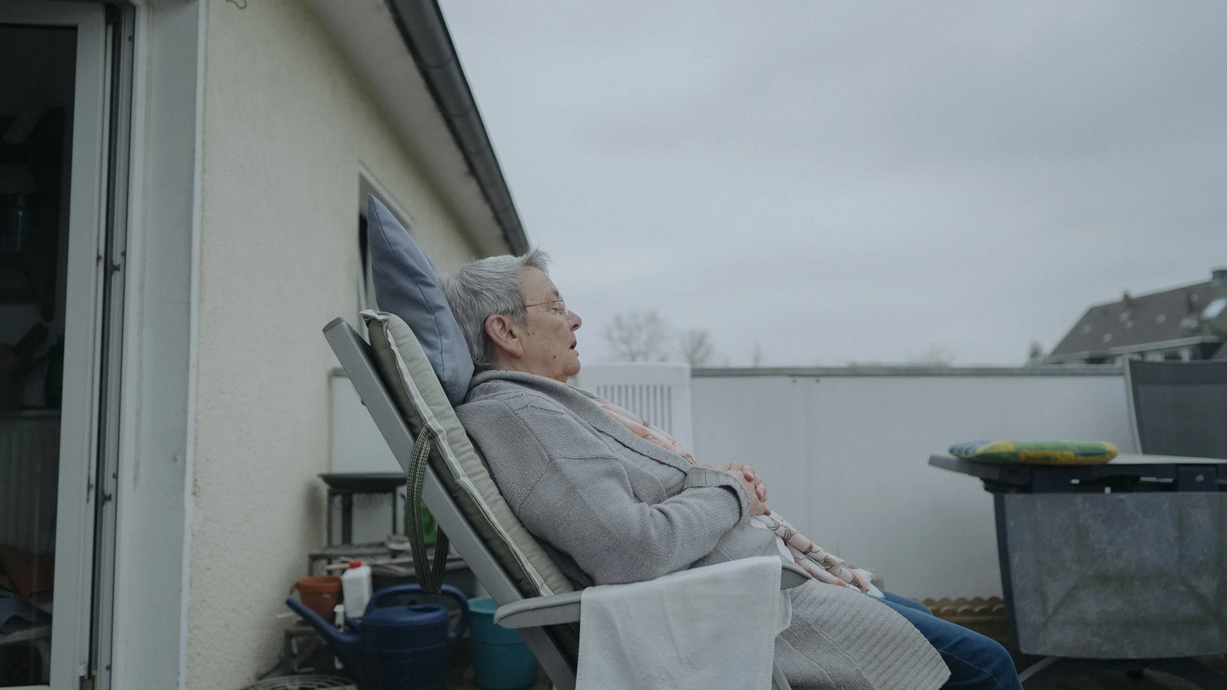 Ältere Frau sitzt entspannt auf einem Balkon, während es draußen bewölkt ist.