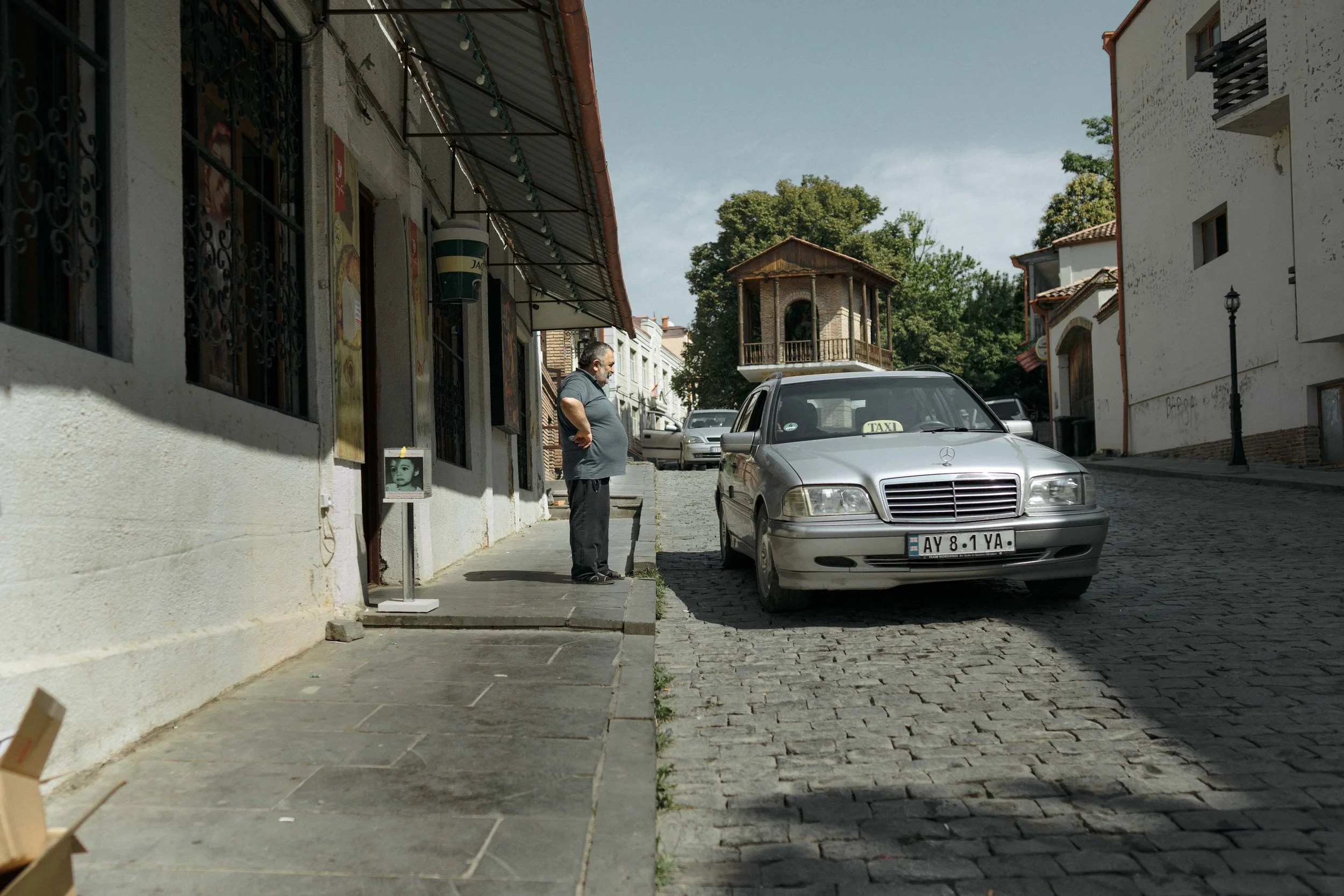 Ein älterer Mann steht auf dem Bürgersteig neben einem grauen Taxi, das auf einer Kopfsteinpflasterstraße parkt, mit einem typischen europäischen Haus im Hintergrund.