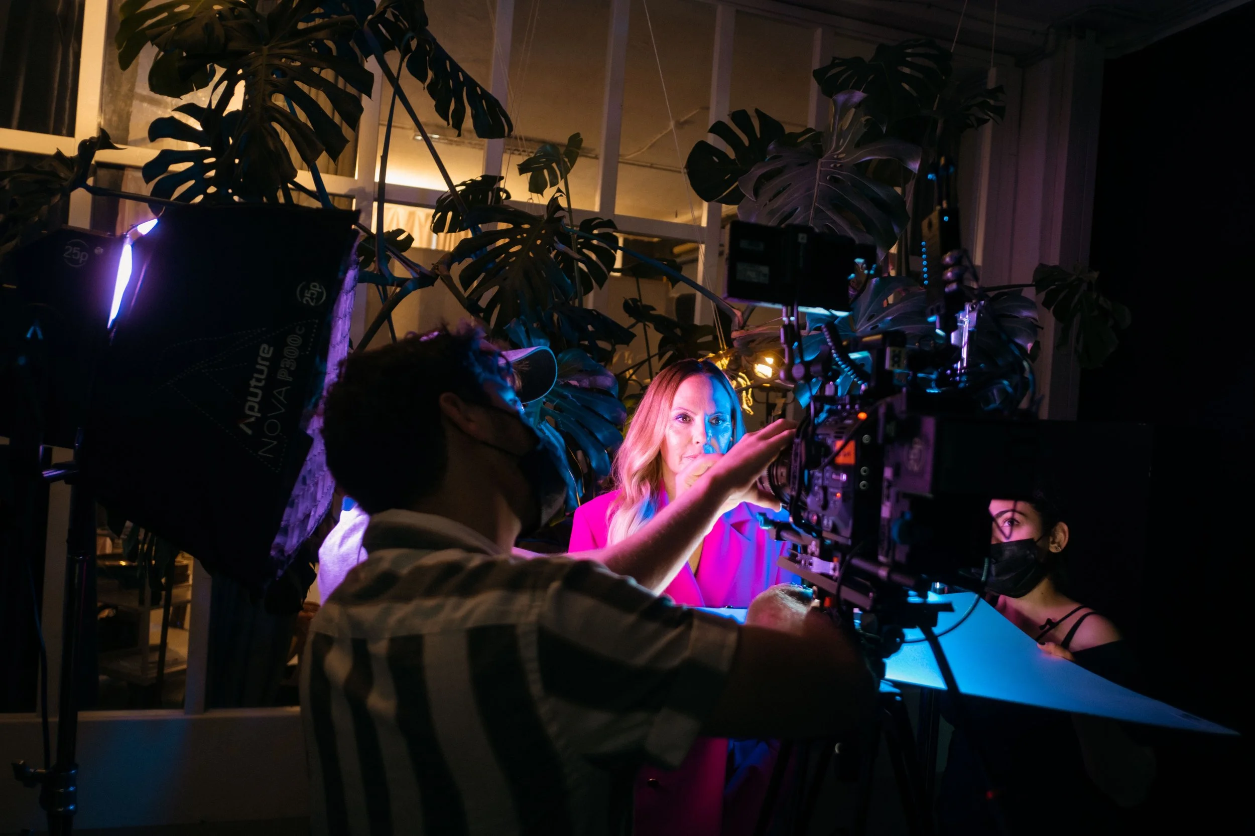 Eine Frau vor einem Kamerastudio mit buntem Licht, umgeben von Pflanzen und einem Mann, der die Kamera bedient, in einem dunklen Raum.