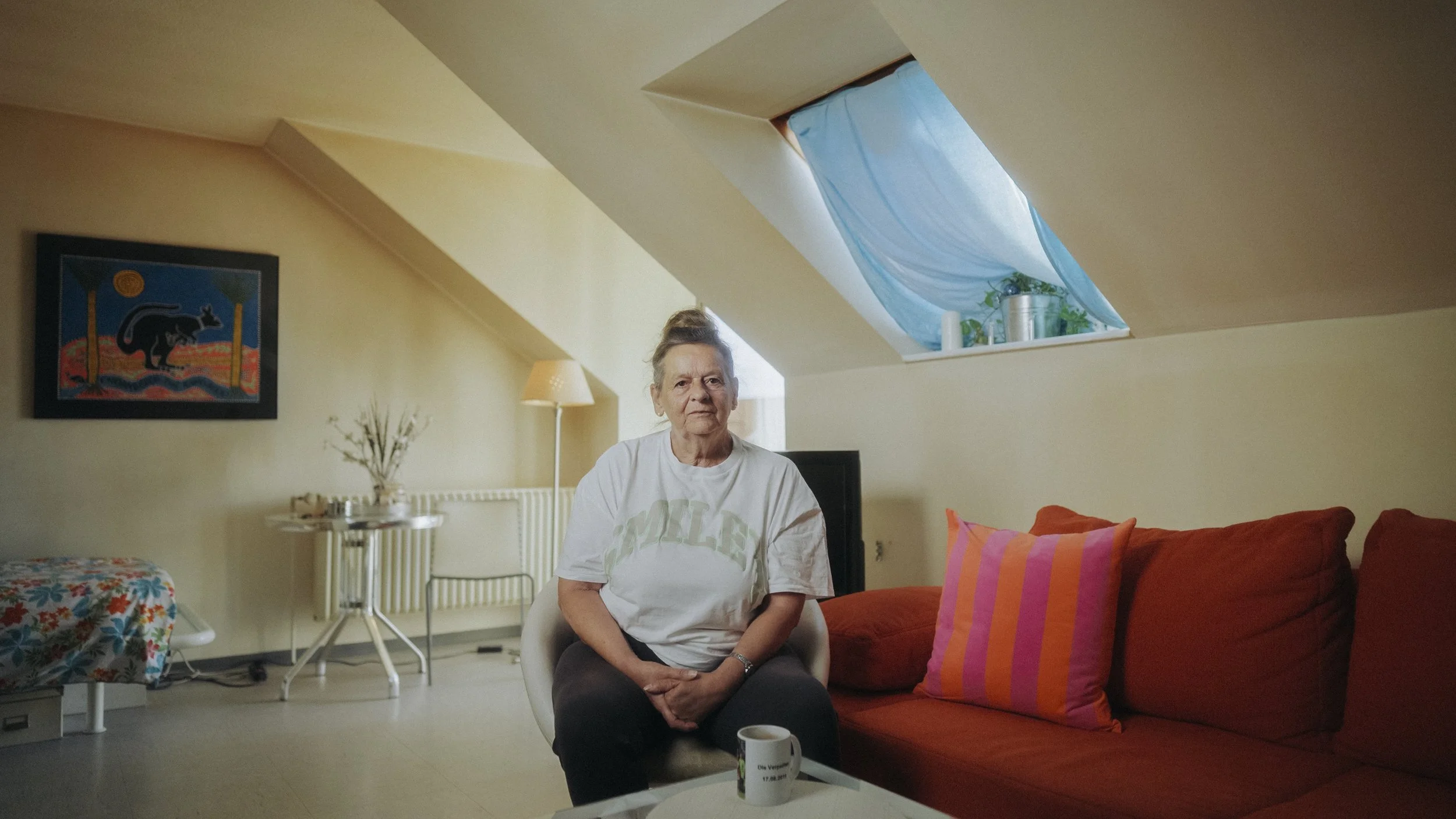 Eine ältere Frau sitzt in einem Wohnzimmer auf einem weißen Stuhl, neben einem roten Sofa mit pink-orangefarbenen Kissen. Im Hintergrund befindet sich eine kleine Lampe, ein Tisch mit Dekoration, und ein Bild an der Wand. Das Zimmer hat ein Dachfenst