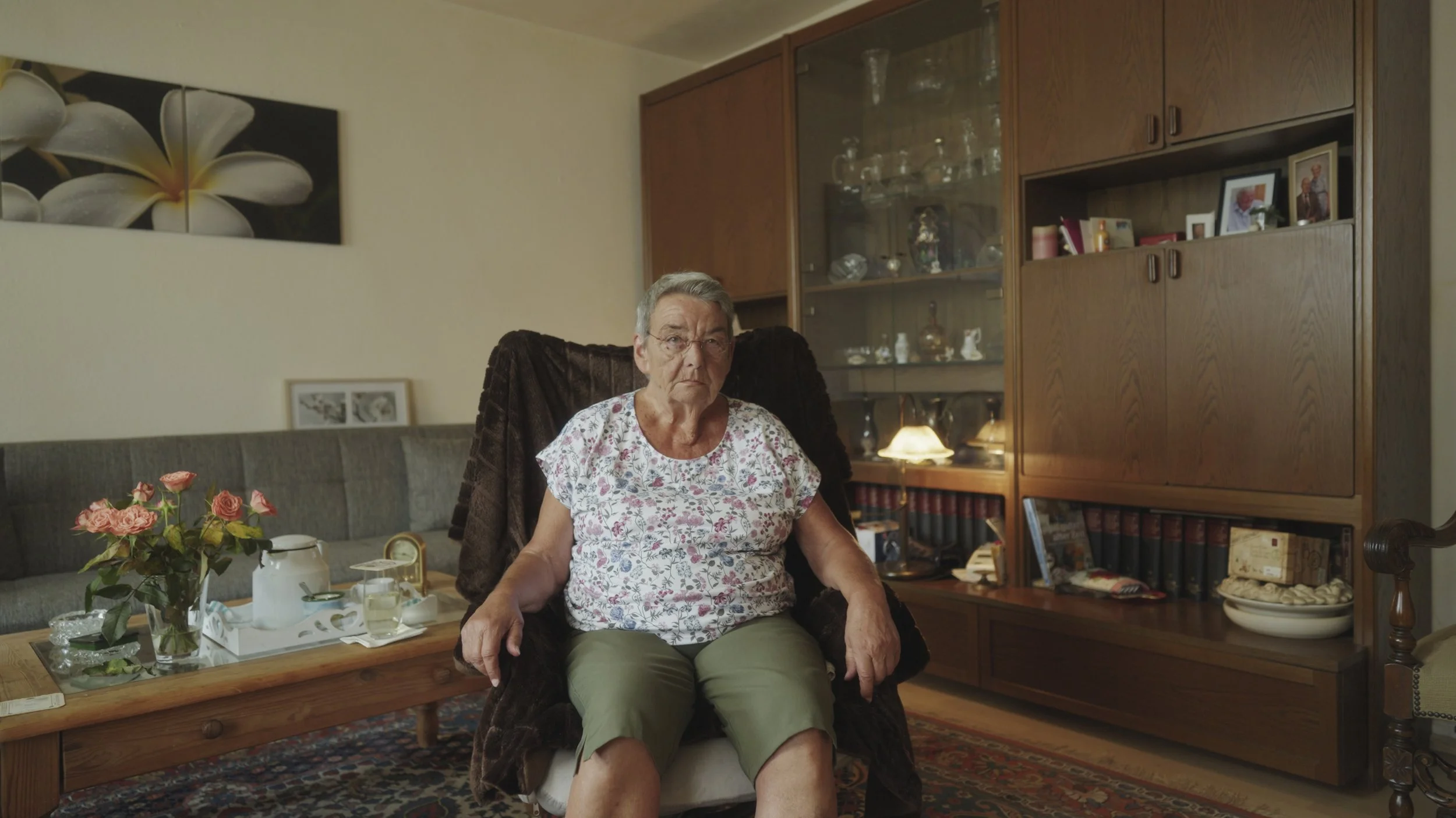 Eine ältere Frau sitzt im Wohnzimmer in einem Sessel, mit einem schwarzen Fellüberwurf. Im Hintergrund befindet sich ein Holschrank mit Glasvitrine und Fotos. Auf dem Couchtisch sind Blumen, eine Teekanne und Gläser.