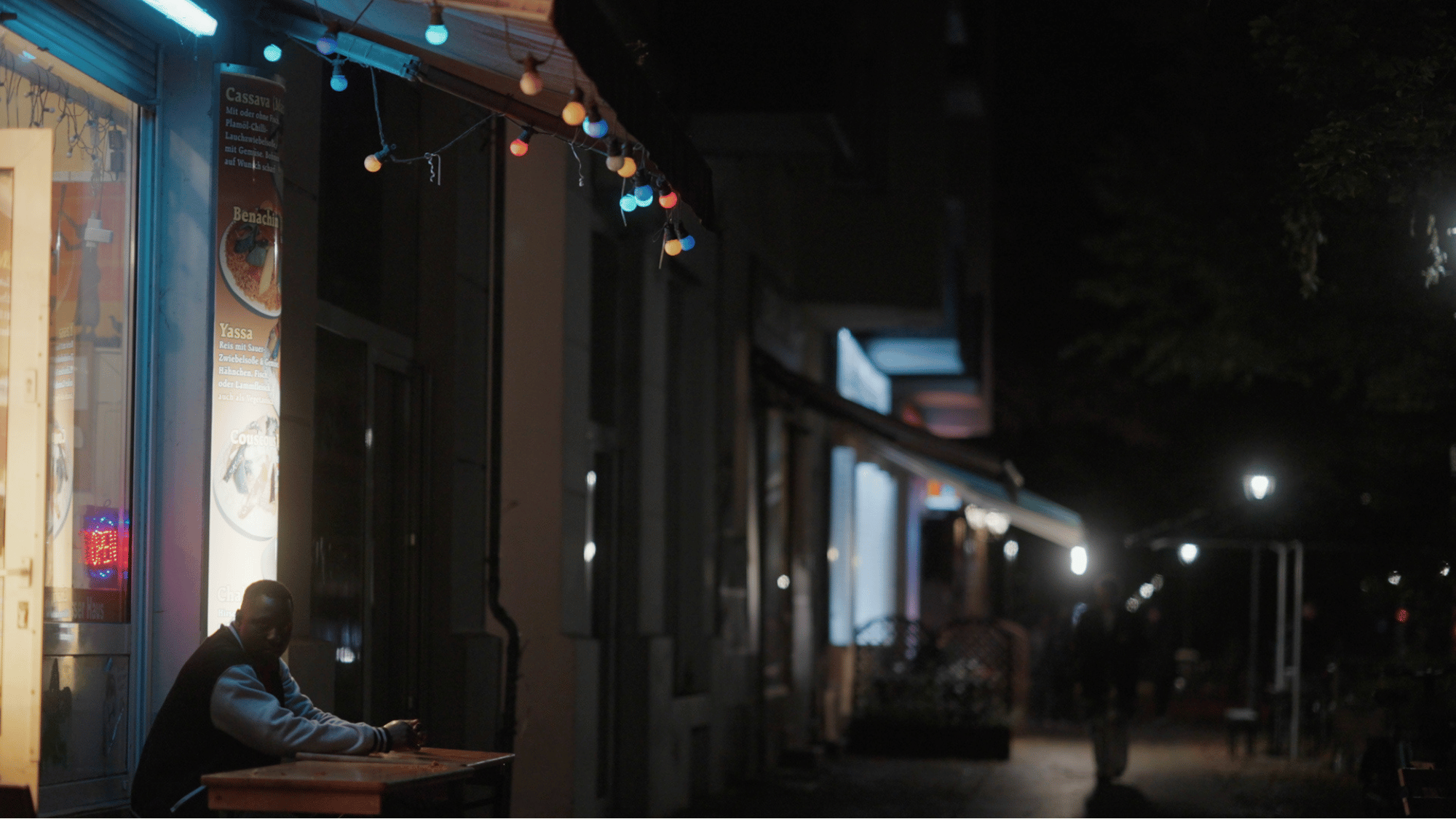 Eine dunkle Straßenszene mit mehreren beleuchteten Geschäften. Ein Mann sitzt an einem Tisch vor einem Geschäft, hinter ihm sind Poster und Lichter. Die Straßen sind kaum beleuchtet, und es gibt nur wenige Fußgänger.