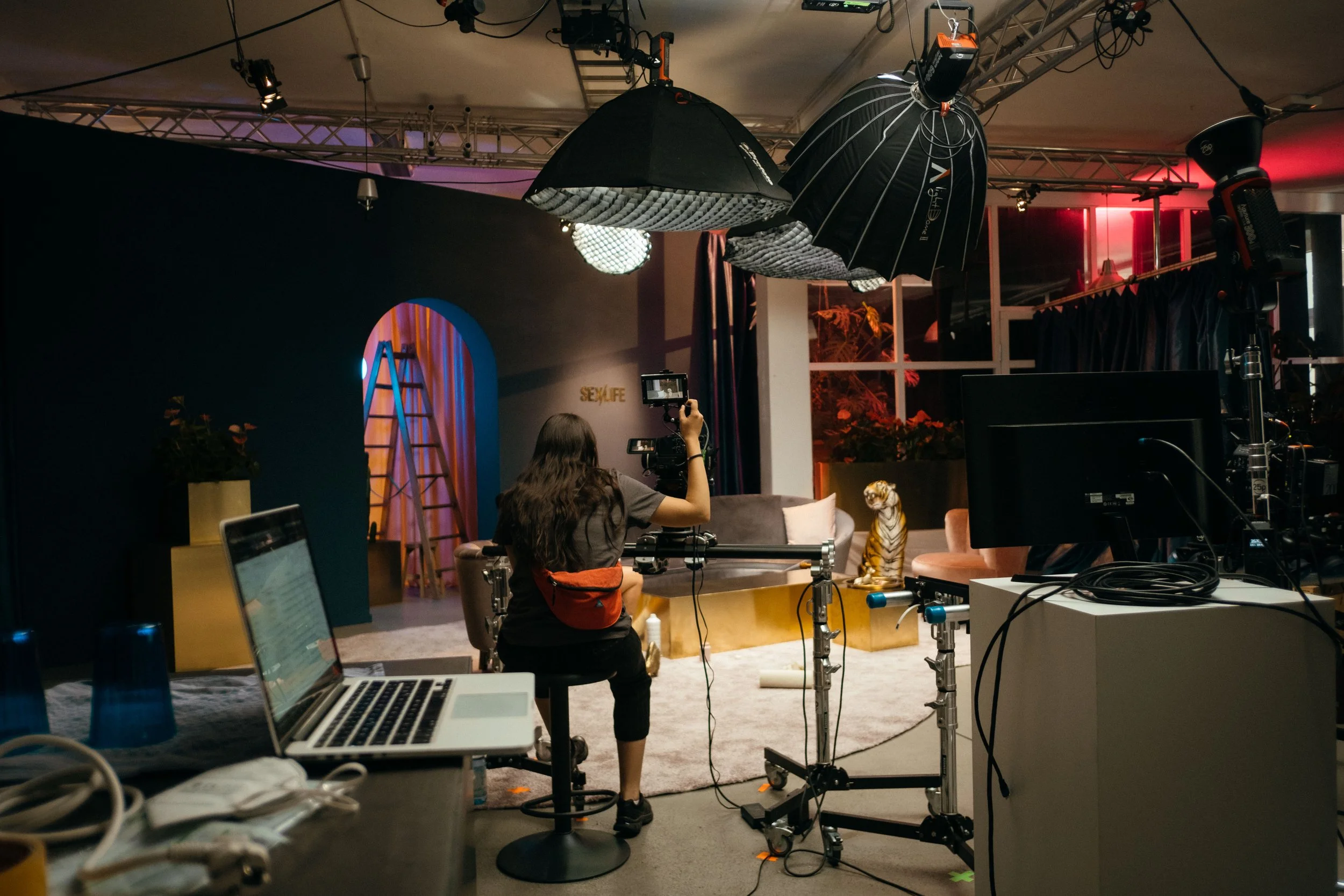 Eine Person sitzt vor einem Video-Set mit professioneller Kamera, Fotografenlicht und Beleuchtung, während sie einen Tiger-Requisite positioniert, und es sind technische Geräte und Kabel im Vordergrund sichtbar.