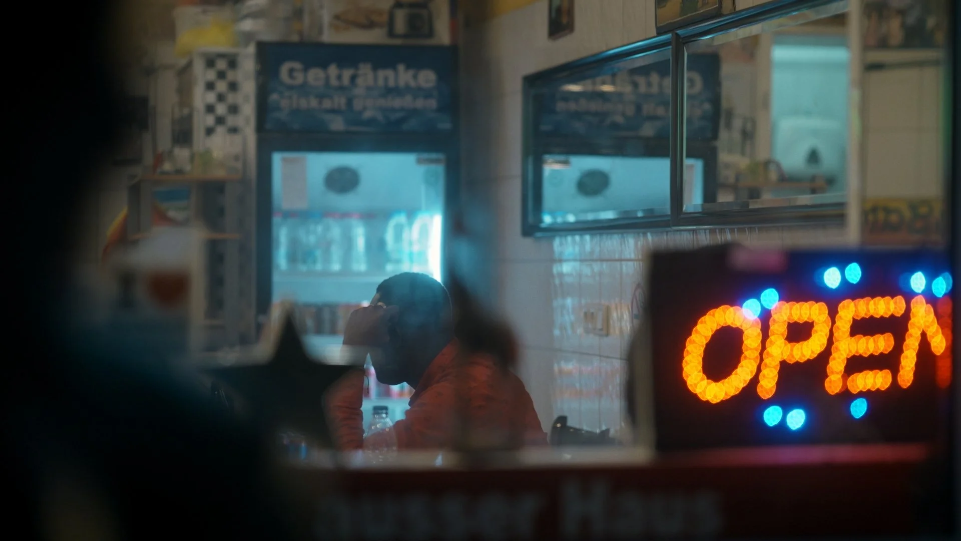 Ein Mensch sitzt in einem kleinen Raum oder einer Cafeteria, sichtbar durch eine Scheibe, die ein leuchtendes 'OPEN'-Schild zeigt. Im Hintergrund sind Getränkeanzeigen und Flaschen sichtbar.