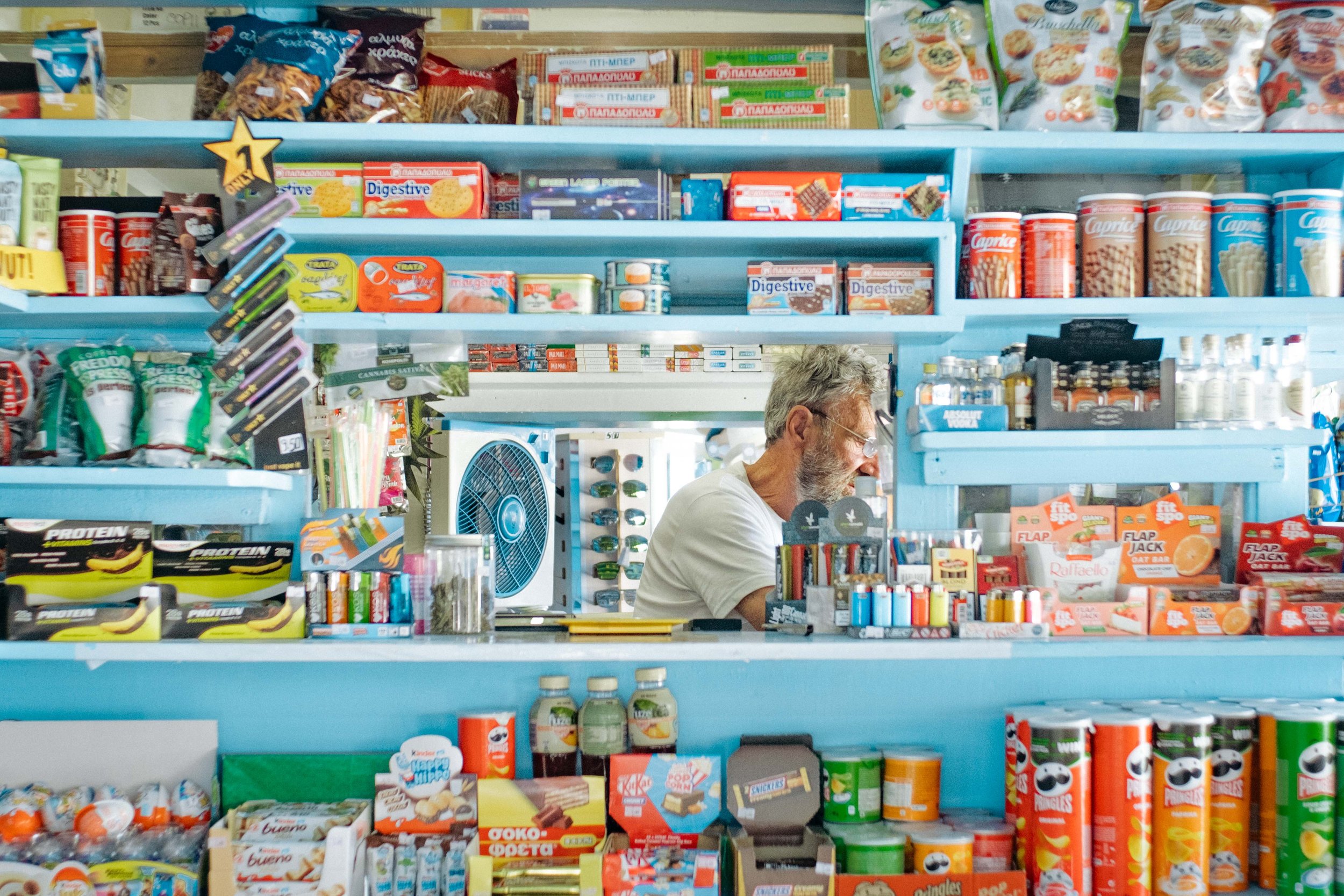 Ein Mann steht hinter Theken in einem Laden, der mit bunten Lebensmitteln und Snacks gefüllt ist.