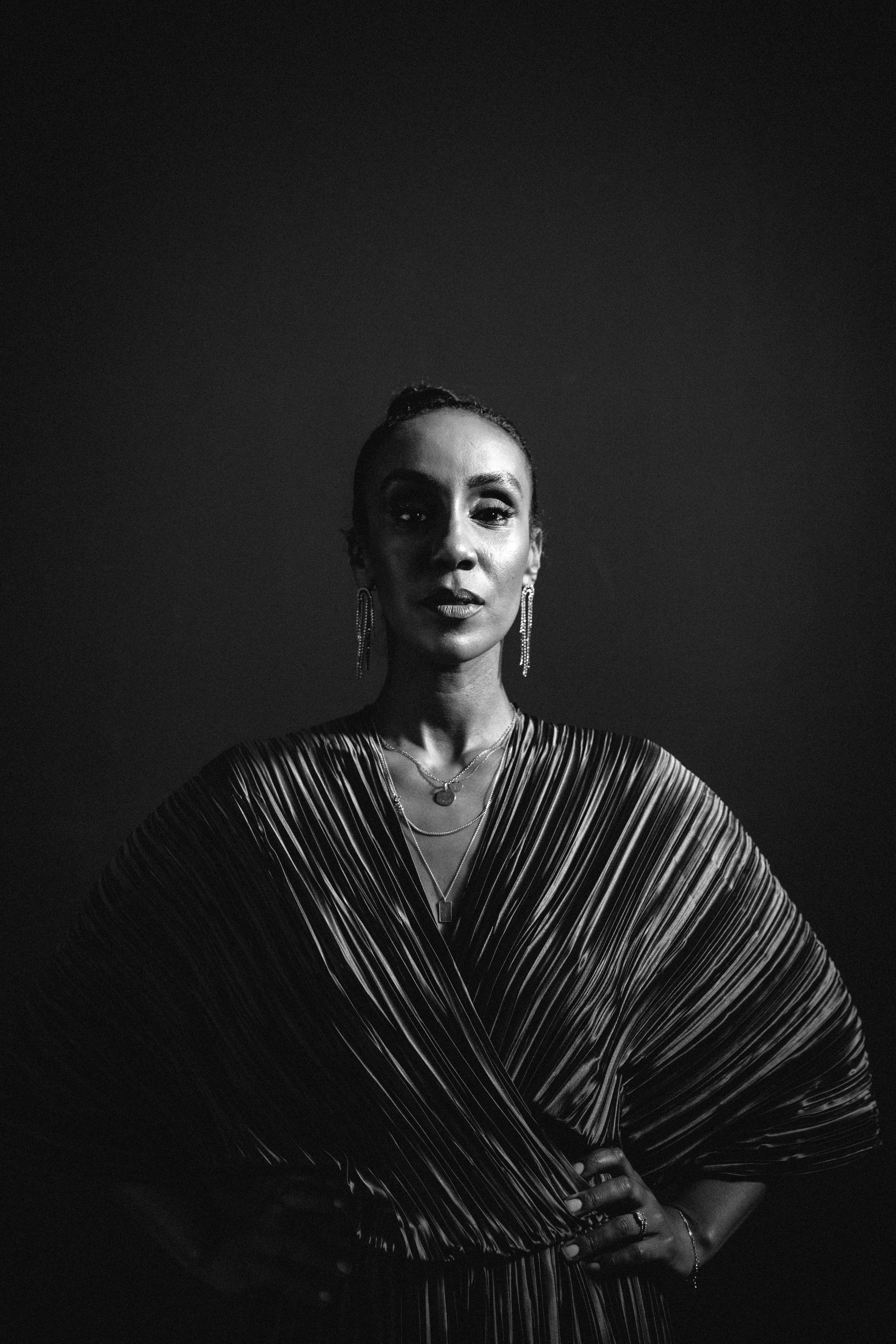 Schwarz-Weiß-Foto einer eleganten Frau, die direkt in die Kamera schaut, mit hervorgehobenen Lippen und auffälligen Ohrringen, stilvoll gekleidet in ein seidiges, nassglänzendes Kleid, mit lockeren Frisur und mehreren Halsketten, vor einem dunklen Hi