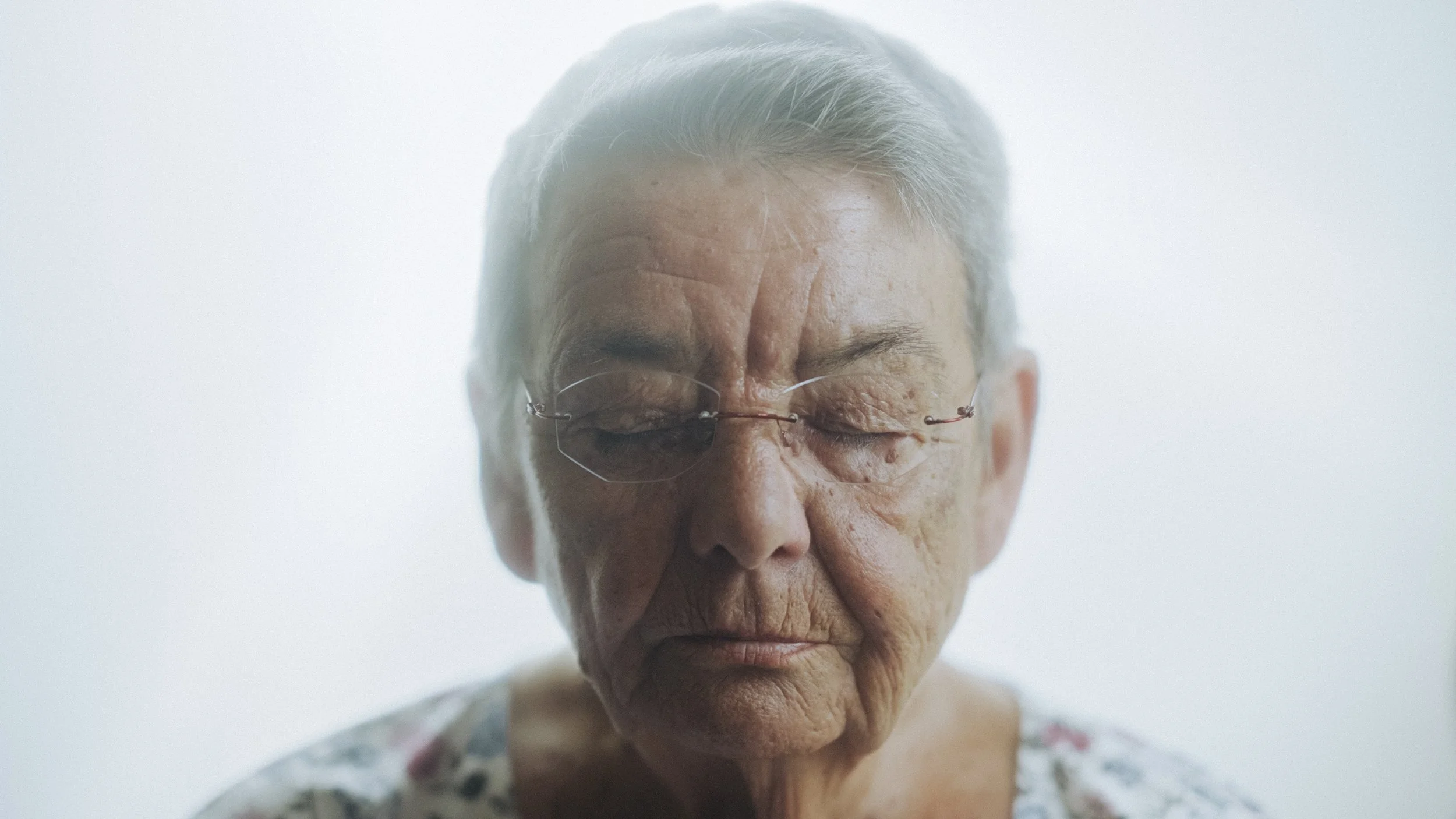 Nahaufnahme einer älteren Frau mit geschlossenen Augen, grauem Haar und Brille, gegen einen hellen, unscharfen Hintergrund.